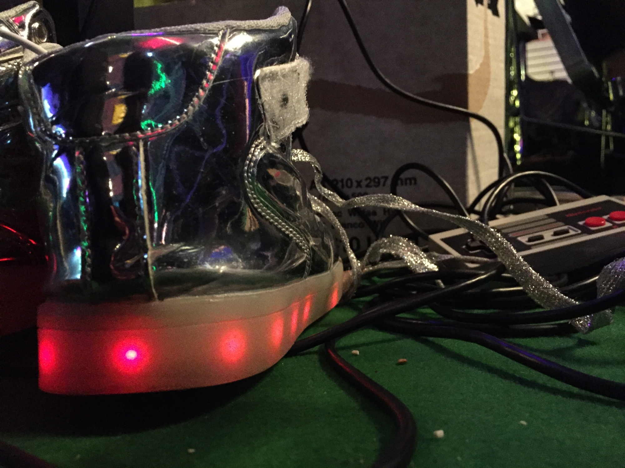 Schuhe mit blinkender Sohle, die gerade zum Laden am USB-Kabel hängen, rechts daneben ein NES-Controller von Nintendo