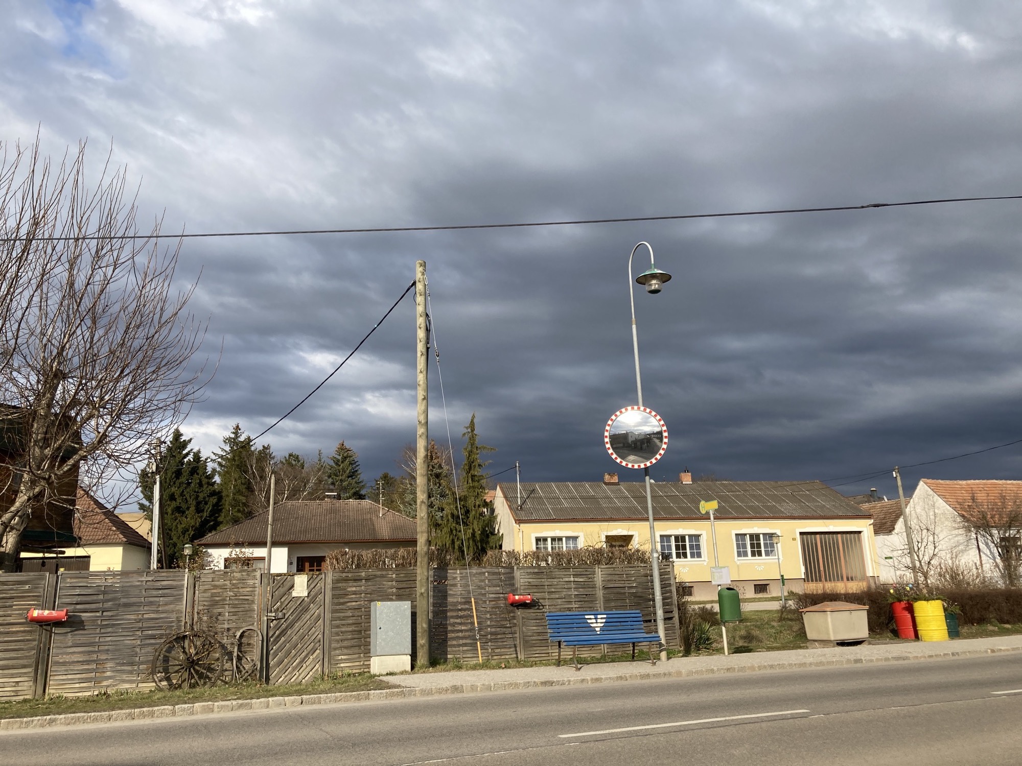 Dorfidylle in Siebenhirten: Busstation mit blauer Bank, alten Feuerlöschern als Blumenkästen, runder Verkehrsspiegel und drei bemalte Tonnen mit Blumen drin