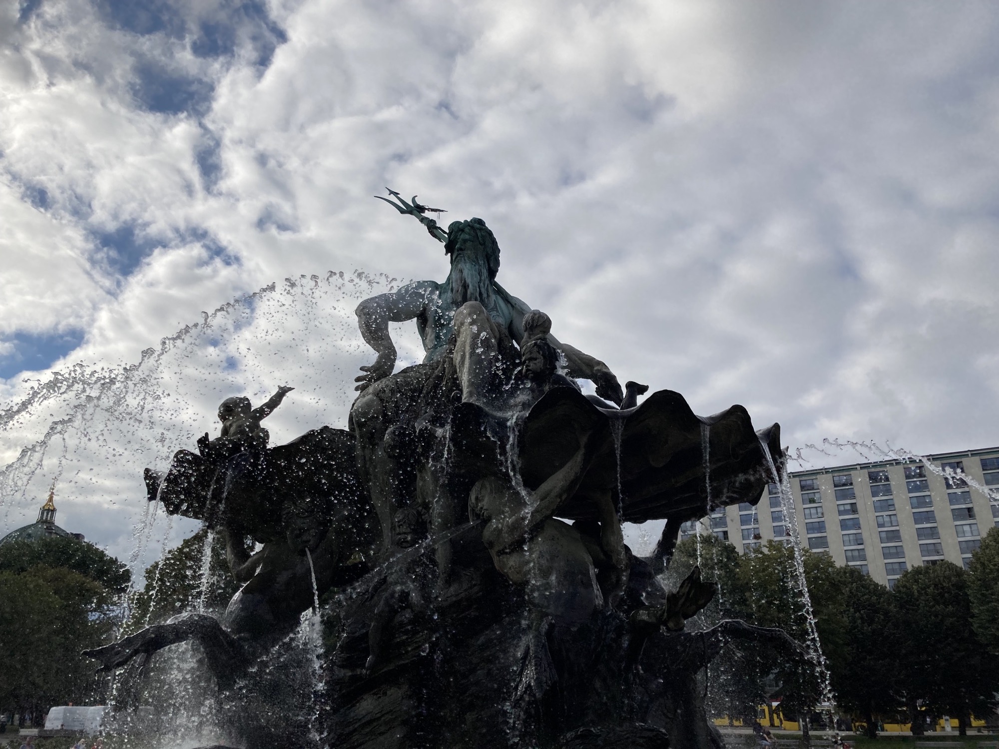 Neptunbrunnen in dramatischer Perspektive, mit fotografisch eingefrorenen Wassertropfen vor bewölktem Himmel