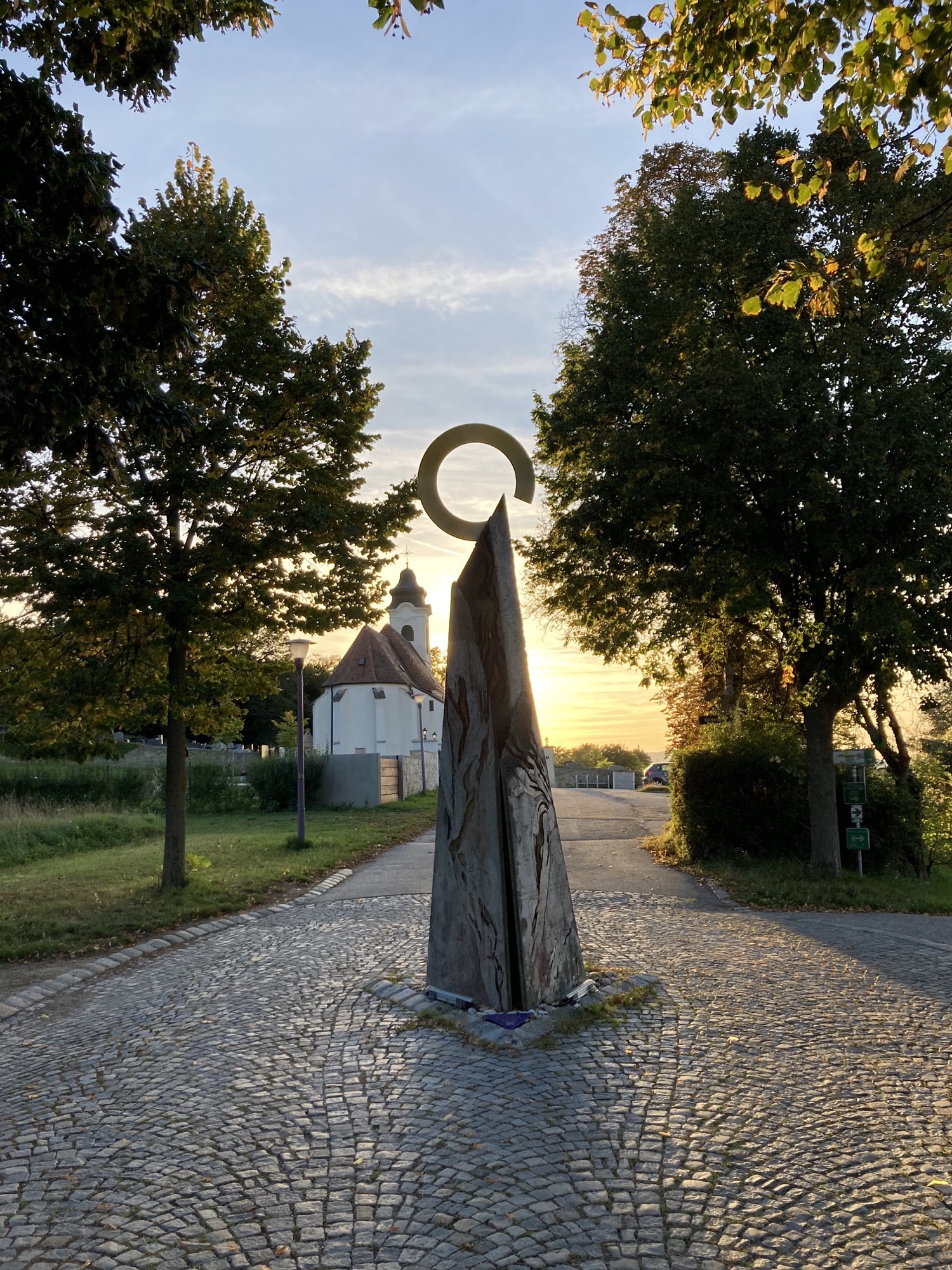 Station 7, eine felsenartige Skulptur, gekrönt von einem nicht geschlossenen Ring, links dahinter Kirche, Friedhof und Abendsonne