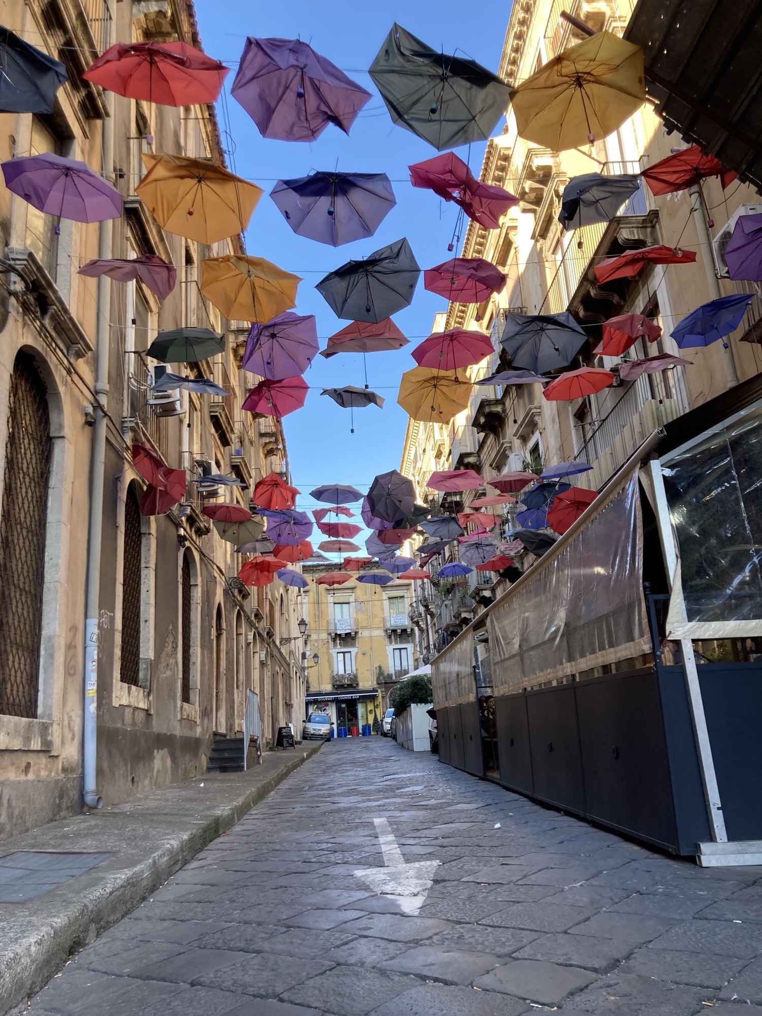 Gasse mit gelben Häusern, auf Höhe des ersten Stocks sind bunte Regenschirme aufgehängt, auf der Straße zeigt ein weißer Pfeil direkt auf die Fotografin