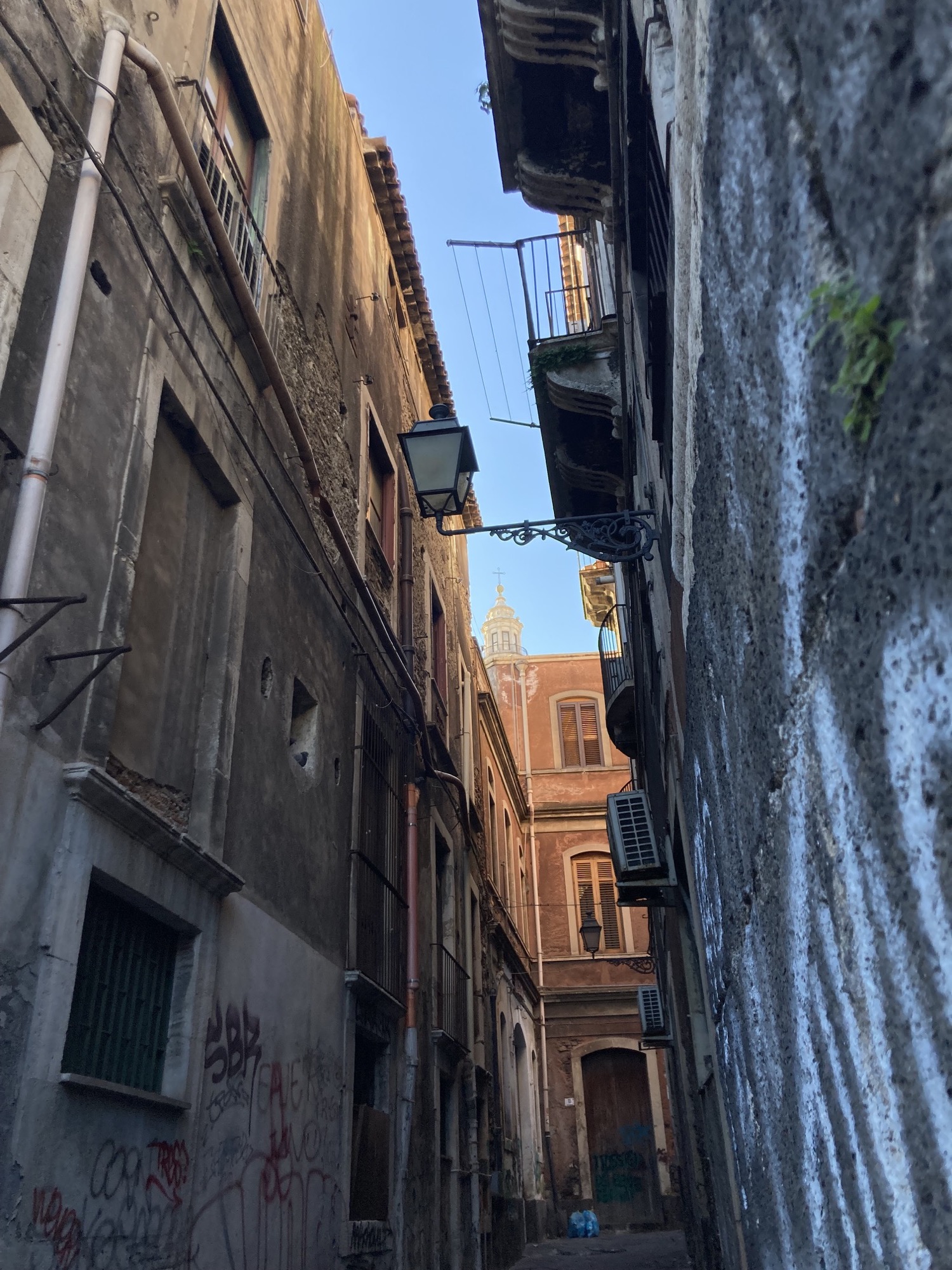sehr schmale Gasse im Schatten mit Balkonen und Klimanlagen, im Hintergrund vor blauem Himmel die Spitze einer Kirche