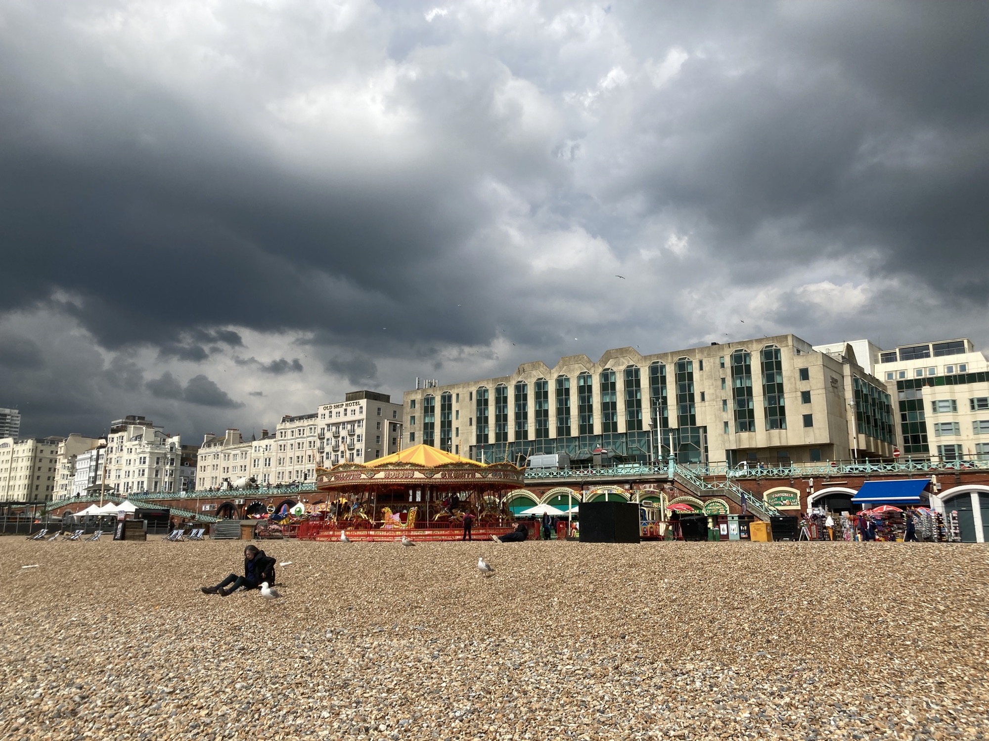 Kieselstrand mit Möwen, im Mittelgrund ein Karussell, dahinter ein imposantes Hotel mit trapezähnlichen Fenstern, im Hintergrund bedrohliche Wolken