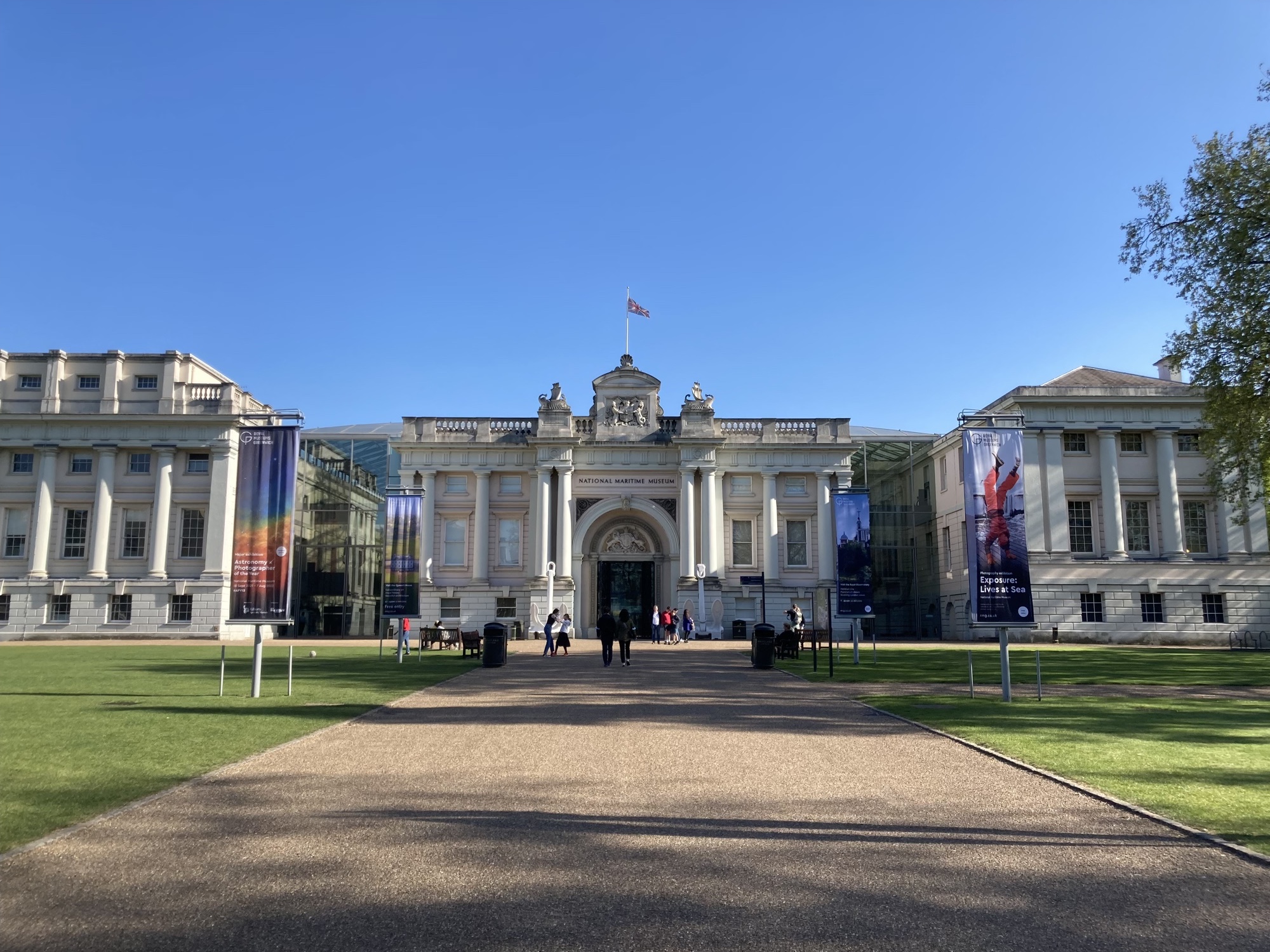 nördlicher Eingang des National Maritime Museums, das weiße Gebäude ist mit Säulen geschmückt und wird von einer Union-Jack-Flagge gekrönt, links und rechts des Weges kündigen Flaggen die aktuellen Ausstellungen an