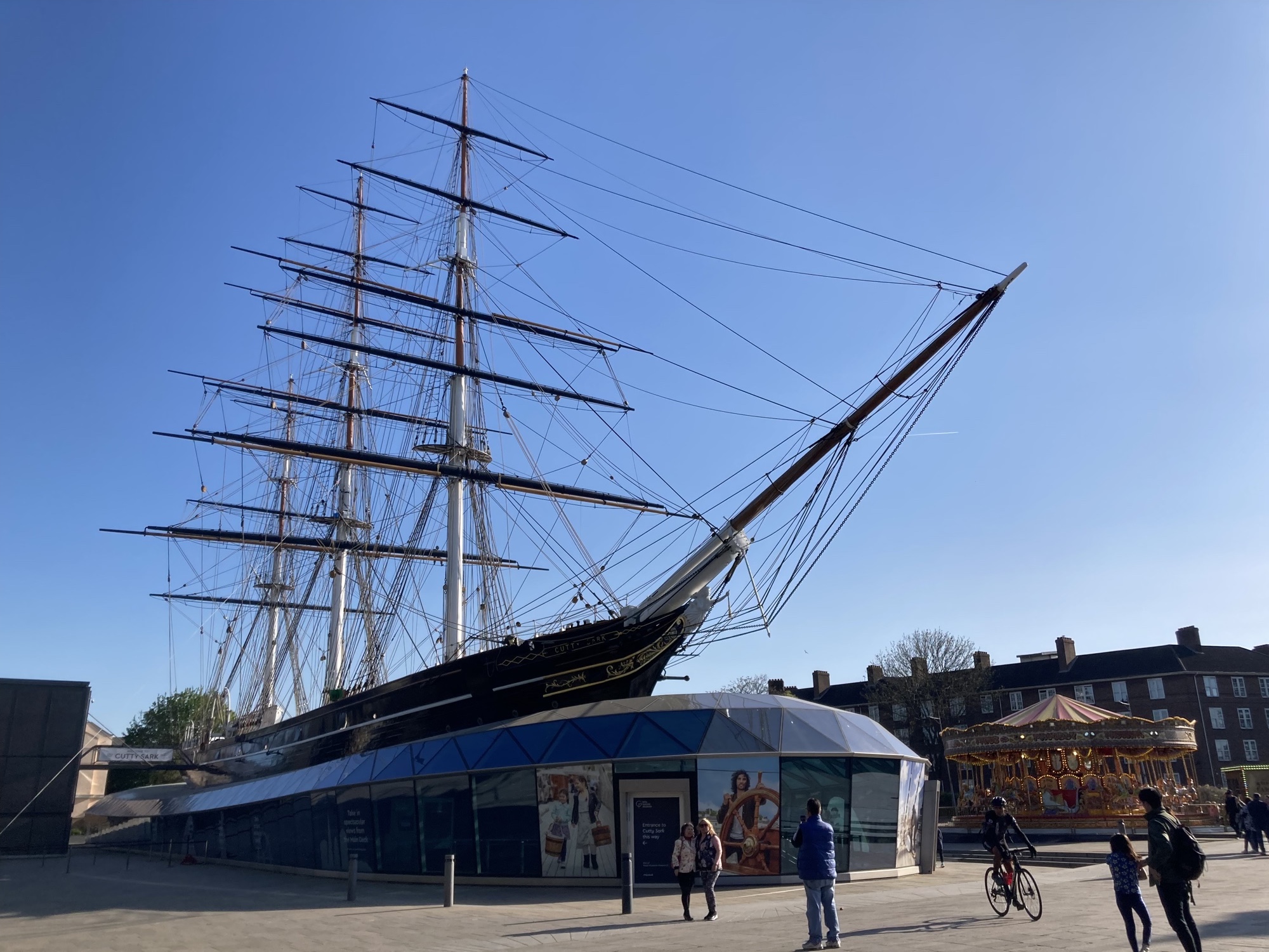 Museumsschiff Cutty Sark, ein Segelschiff mit drei Masten, das auf einem Glasgebäude ruht, rechts hinter diesem Glasgebäude ist ein Karussell zu sehen