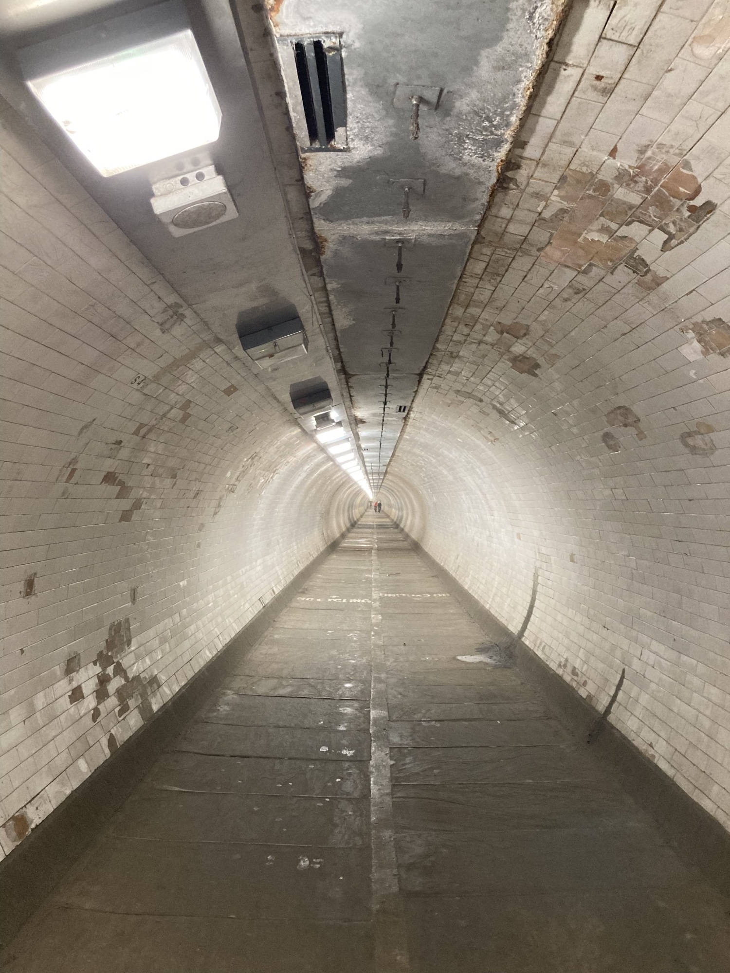 Tunnelblick in den Greenwich Foot Tunnel, der Boden besteht aus rechteckigen grauen Steinen, die Wände der Tunnelröhre sind mit weißen Fliesen ausgekleidet, die an vielen Stellen deutliche Abnutzungserscheinungen aufweisen