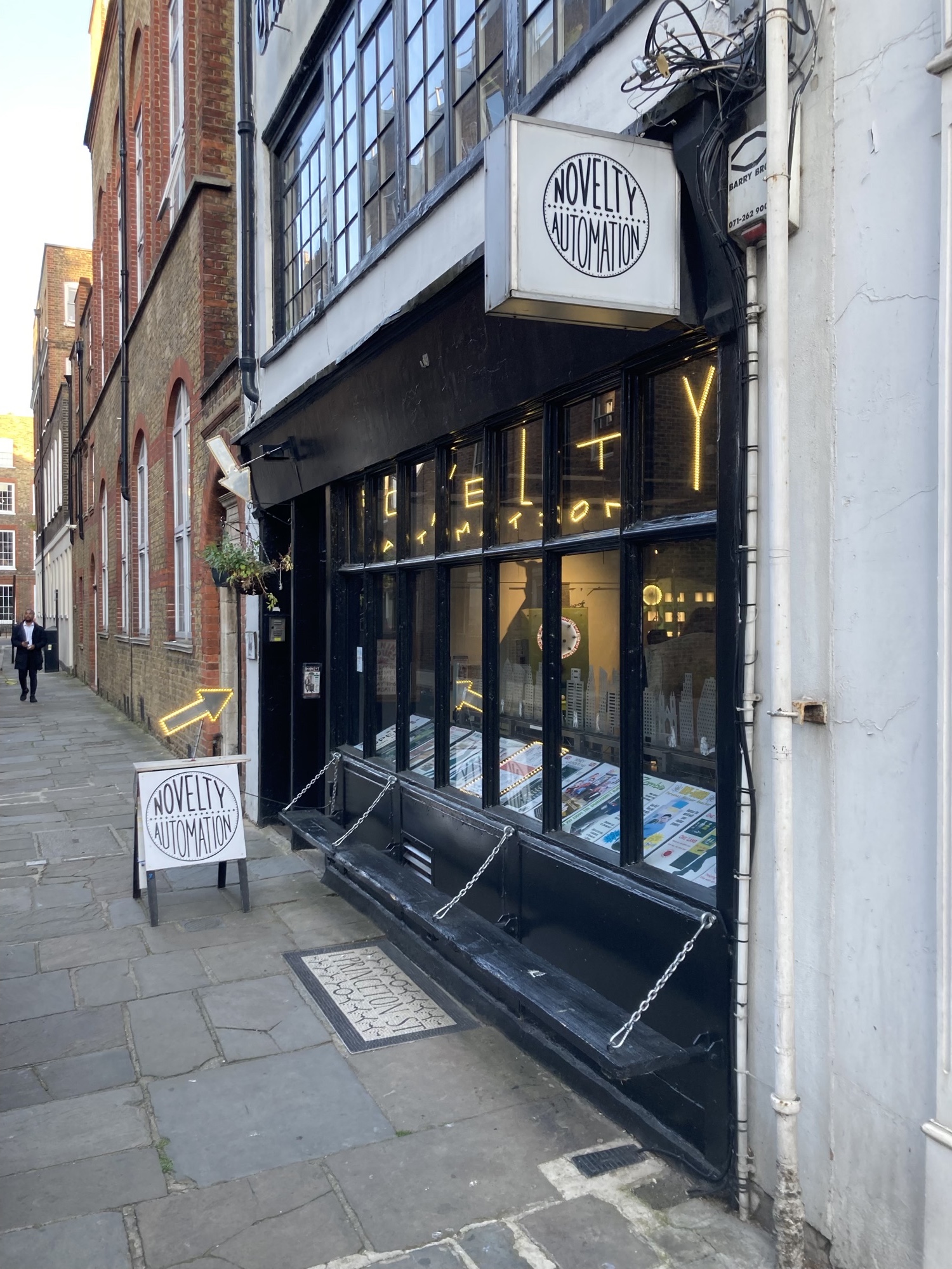 Gebäudefront des Novelty Automation, schwarze Holzverkleidung rund um die Fenster, eine aufklappbare Sitzbank außen, vor der Tür ein Schild mit einem leuchtenten, sich hin und her bewegenden Pfeil