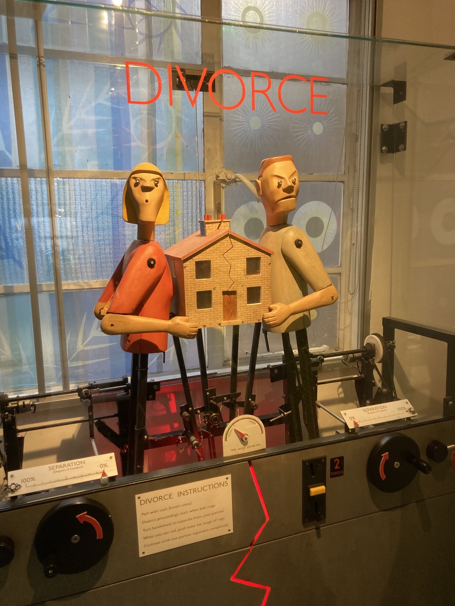 Divorce Automat, in einem Glaskasten befinden sich zwei Puppen, die als männlich und weiblich zu verstehen sind, jede Puppe hält eine Seite eines Hauses, beide haben einen grimmigen Gesichtsausdruck, außen am Kasten befinden sich zwei Kurbeln, an denen gedreht werden muss, um den Automaten zu bedienen