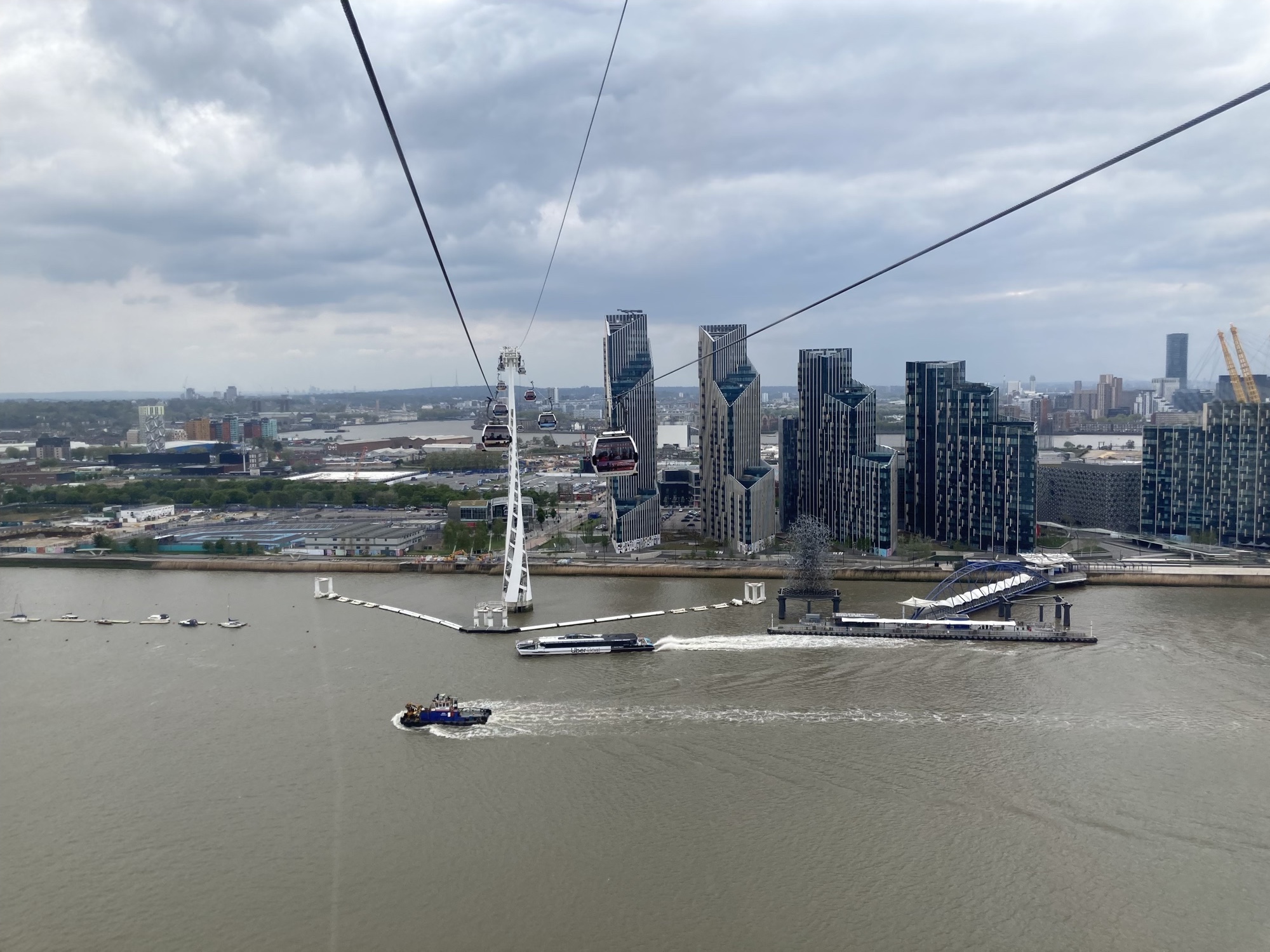 Blick aus der Seilbahn auf die Halbinsel North Greenwich, mittig Seilbahn mit Gondeln und Wolkenkratzer mit Glasfronten, unten die Themse, wo gerade ein Boot die Anlegestation verlässt