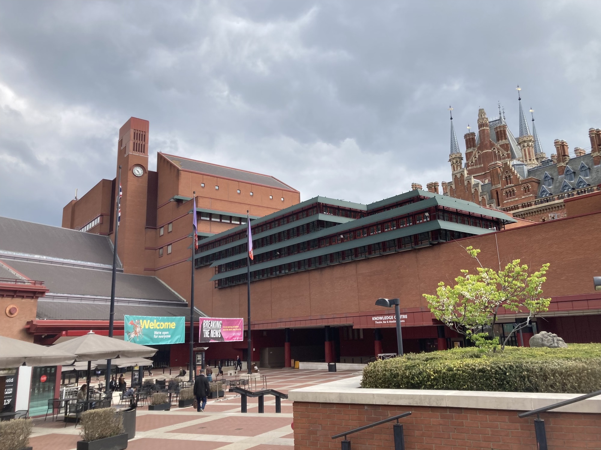 Blick auf den Vorplatz und das Gebäude der British Library, roter Steinbau mit Ausstellungsankündigungen und Fahnen, hinter dem Gebäude sind die Spitzen des Bahnhofs King's Cross St. Pancras zu sehen