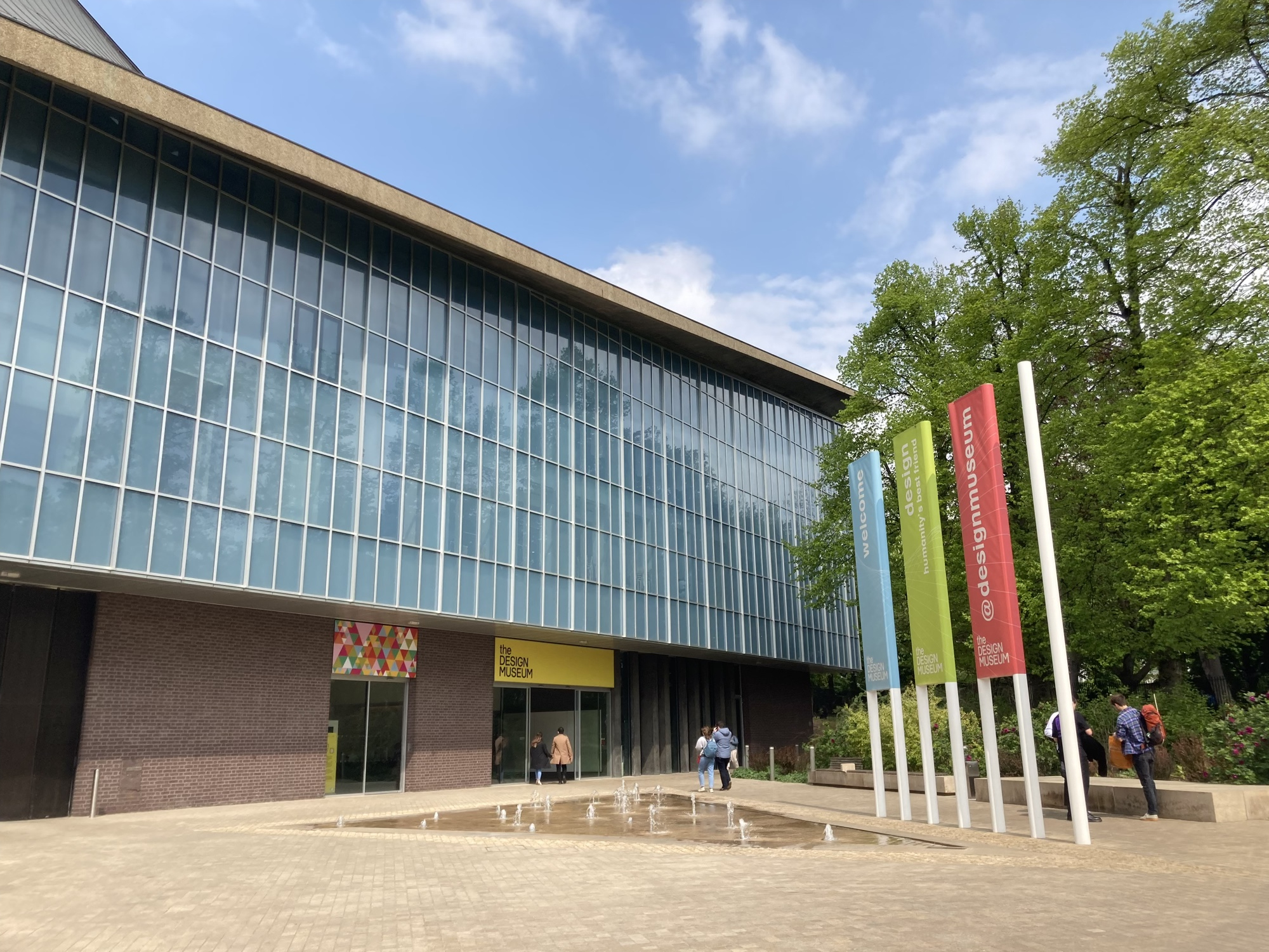 Eingangsbereich des Design Museums in London von außen, eine imposante schräge Glasfront, drei Flaggen in unterschiedlichen Farben, ein bodenebener Brunnen mit mehreren Springquellen