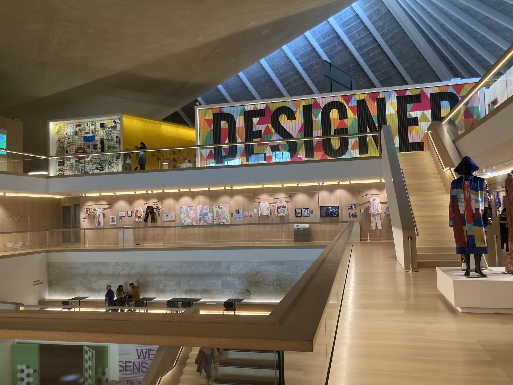 Blick durch den offenen Foyerraum des Design Museums, rechts oben eine Textwand mit dem Schriftzug „Designer“, von dort führt eine Treppe ein Stockwerk tiefer, wo Kleidung aus der Modeausstellung zu sehen ist