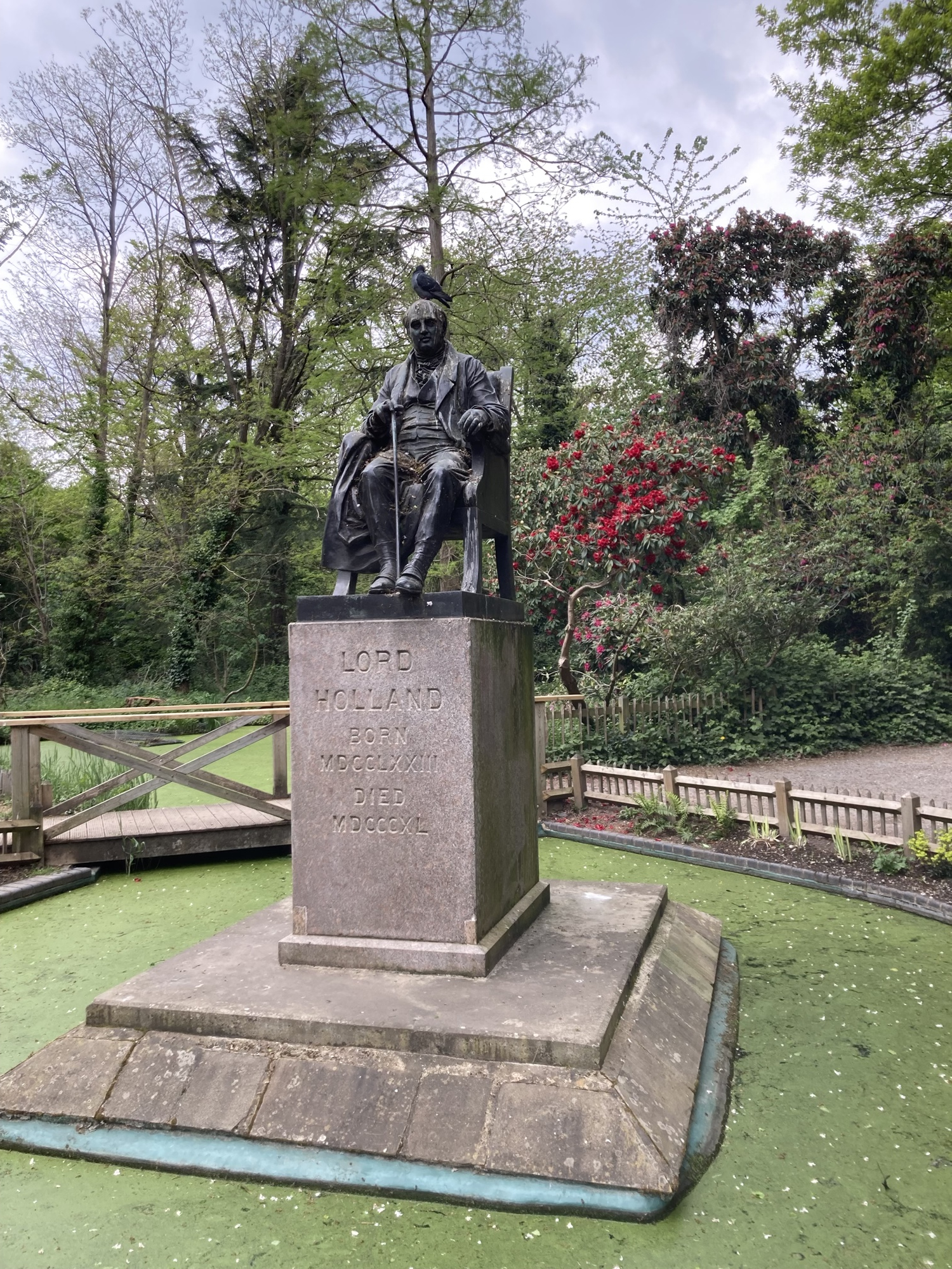 auf einem Sockel inmitten eines von Algen bedeckten Teichs sitzt die Statue von Lord Holland, auf seinem Kopf hat es sich eine Taube gemütlich gemacht