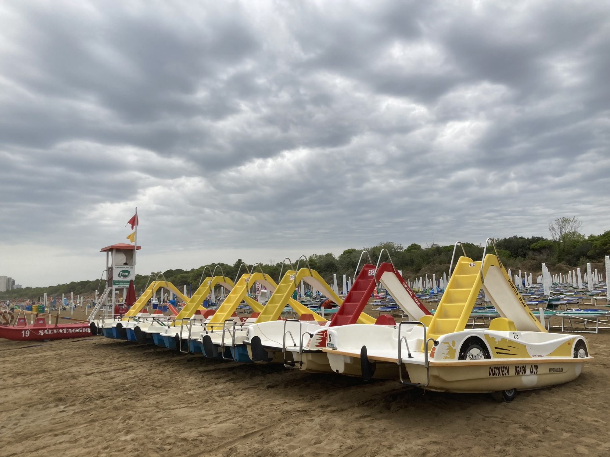 Strand von Lignano, im Vordergrund Tretbotte mit Rutschen darauf auf den Strand gezogen, links davon ein Turm und ein Rettungsboot, dahinter zusammengeklappte Schirme und Strandliegen