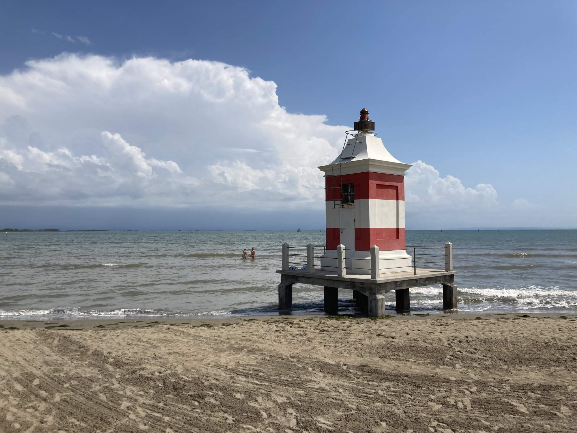 kleiner Leuchtturm am Strand von Lignano, waagrecht rot-weiß gestreift, auf Pfeilern im niedrigen Wasser in den Wellen stehend