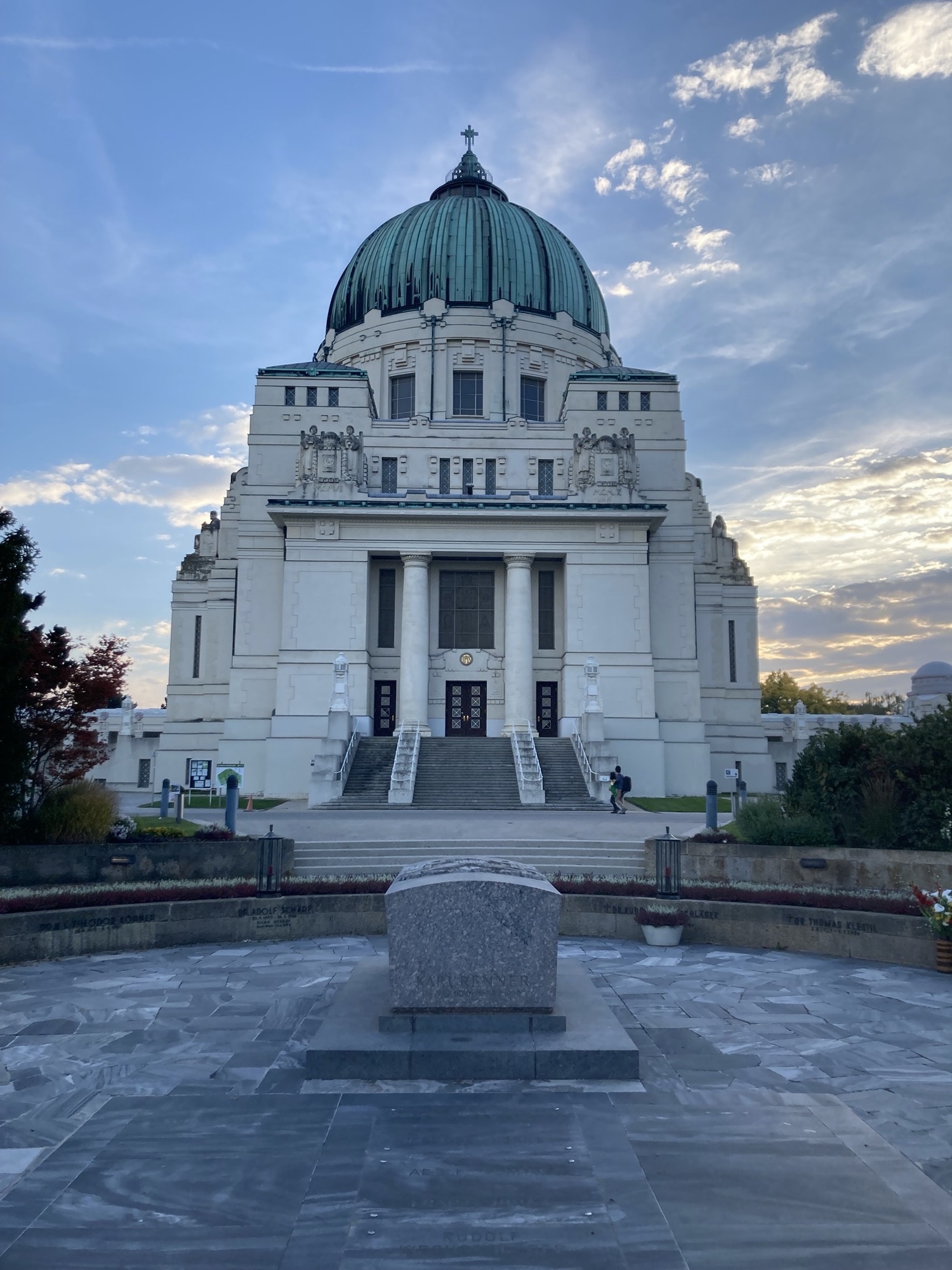 Frontalansicht der Friedhofskirche zum heiligen Karl Borromäus, im Vordergrund die Präsidentengruft, die weiße Kirche mit der grünlichen Kuppel hebt sich im Abendlicht deutlich vom leicht bewölkten Himmel ab