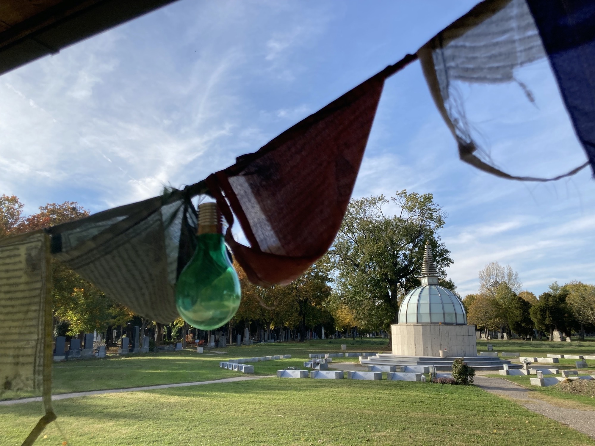 buddhistischer Friedhof Wien, im Hintergrund ein Stupa um ringt von Steinen auf dem Boden in Form eines Rades mit acht Speichen, im Vordergrund etwas unscharf eine Schnur mit Gebetsfahnen und einer grünen Glühbirne