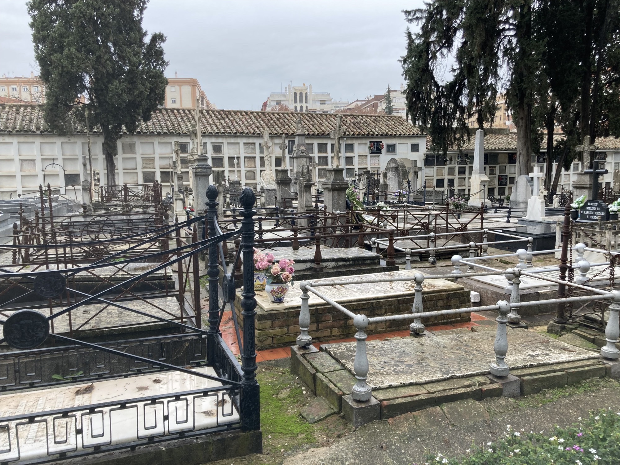 Friedhof, im Vordergrund alte Erdgräber, die von Gittern umspannt und von Kreuzen gekrönt sind, im Hintergrund eine Wand mit Marmortafeln, auf denen die Namen von Verstorbenen verzeichnet sind