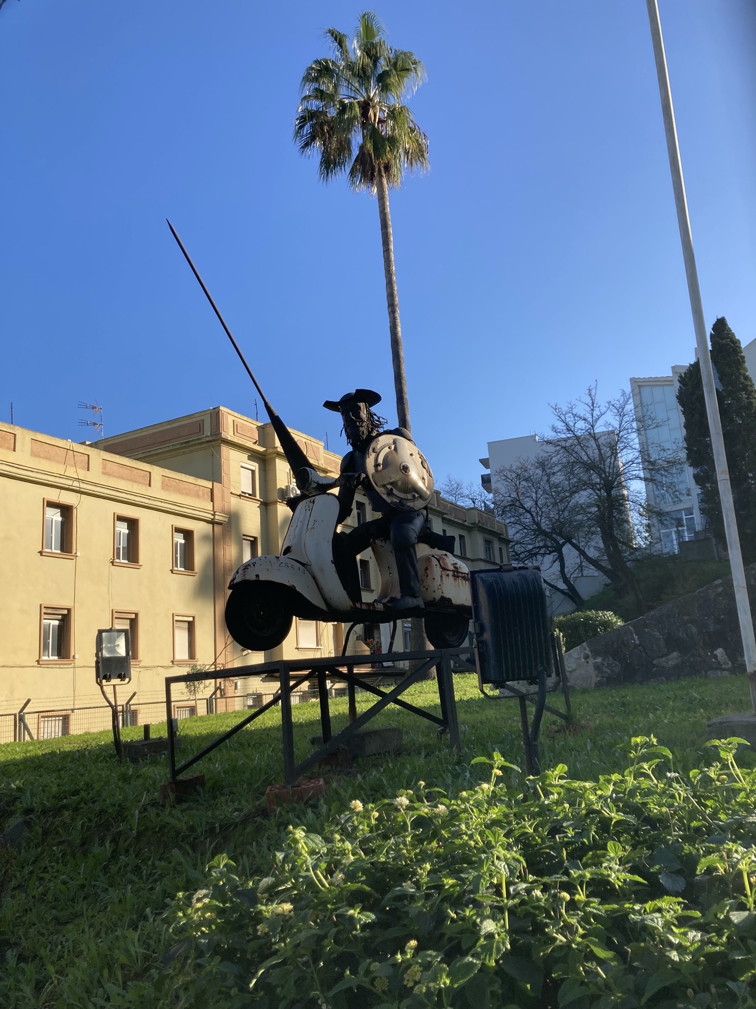eine Skulptur in einem Garten, die Don Quijote de la Mancha darstellt, er hält eine Lanze und einen Schild in den Händen und „reitet“ auf einem weißen Mofa