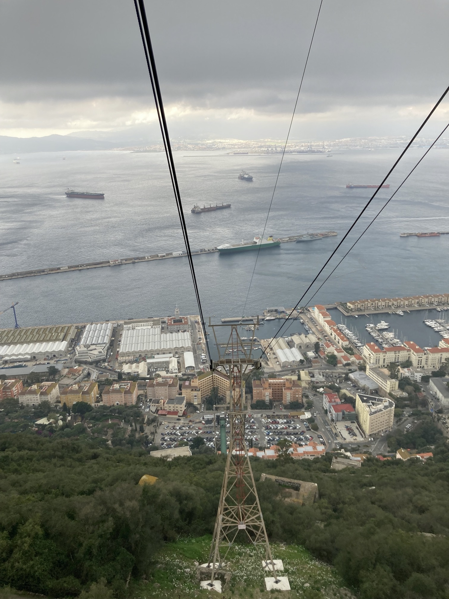 Ausblick aus der Kabine der Seilbahn auf einen Pfosten der Seilbahn, ebenerdig ist die Bodenstation und der dazugehörige Parkplatz zu sehen, dahinter fünf Frachtschiffe, die die Bucht kreuzen und im Hintergrund der Containerhafen und die Stadt Algeciras