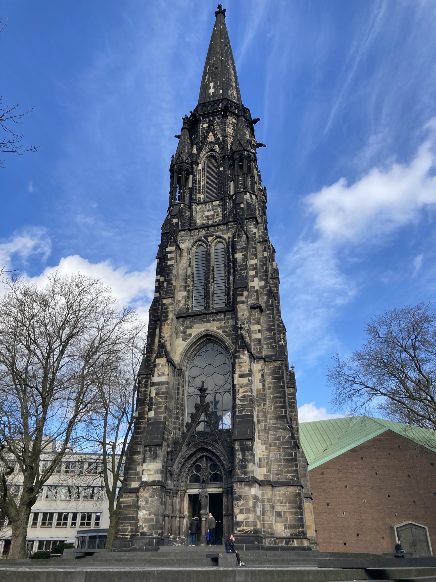 Christuskirche (Kirche der Kulturen), ein schlanker, sich nach oben verjüngender Kirchturm aus Ziegeln im Farbspektrum ocker bis dunkelbraun mit einem Spitzbogen und einem transparenten Rosenfenster über dem Portal