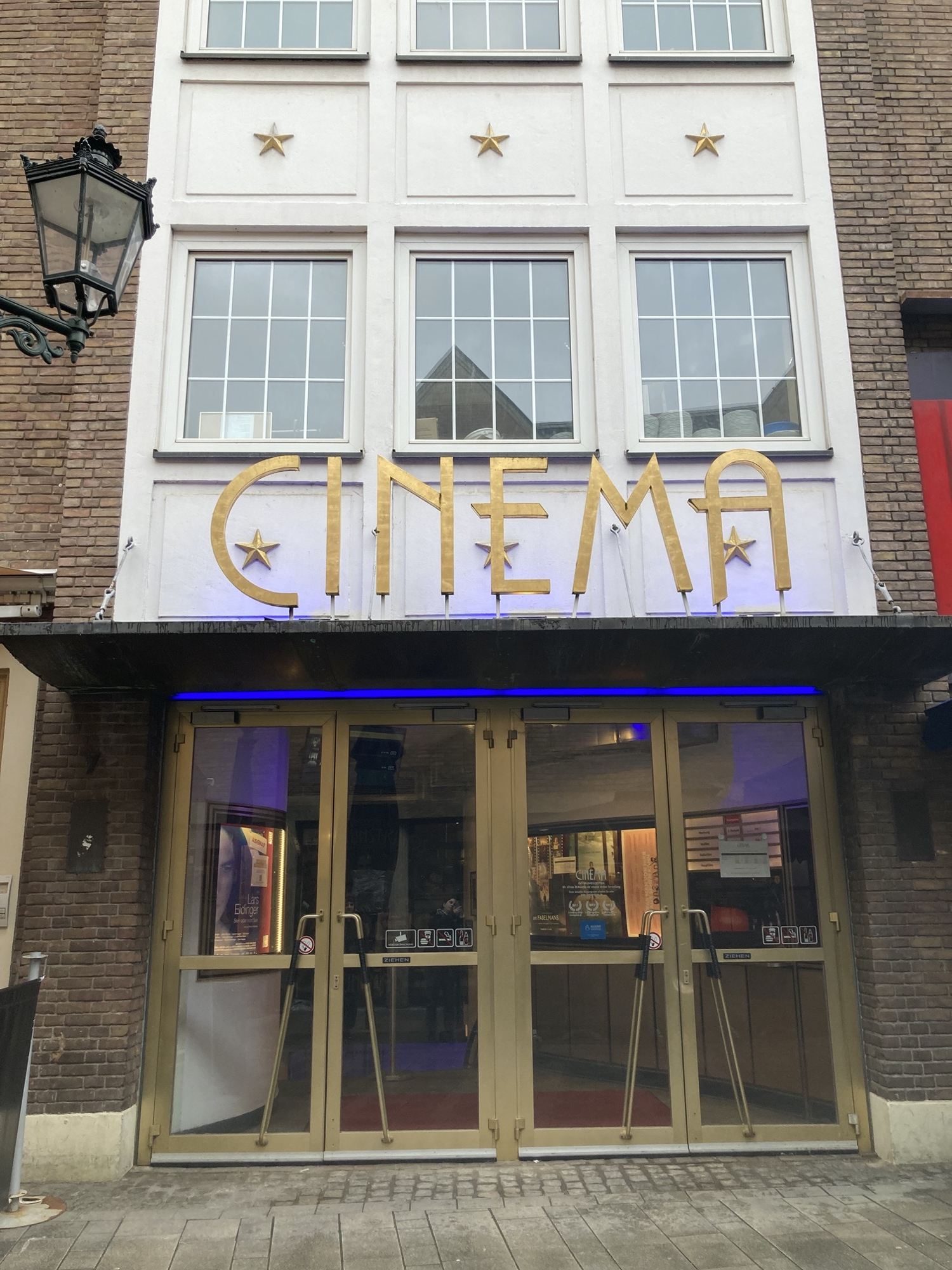 Eingang eines altmodischen Kinos, hinter zwei Glas-Flügel-Türen ist die Kasse des Kinos zu sehen, über dem Eingang der Schriftzug „CINEMA“ in einer schlanken, eckigen Schriftart, dahinter ist ein Teil des Gebäudes weiß verkleidet, zwischen den Fenstern sind goldene Sterne montiert