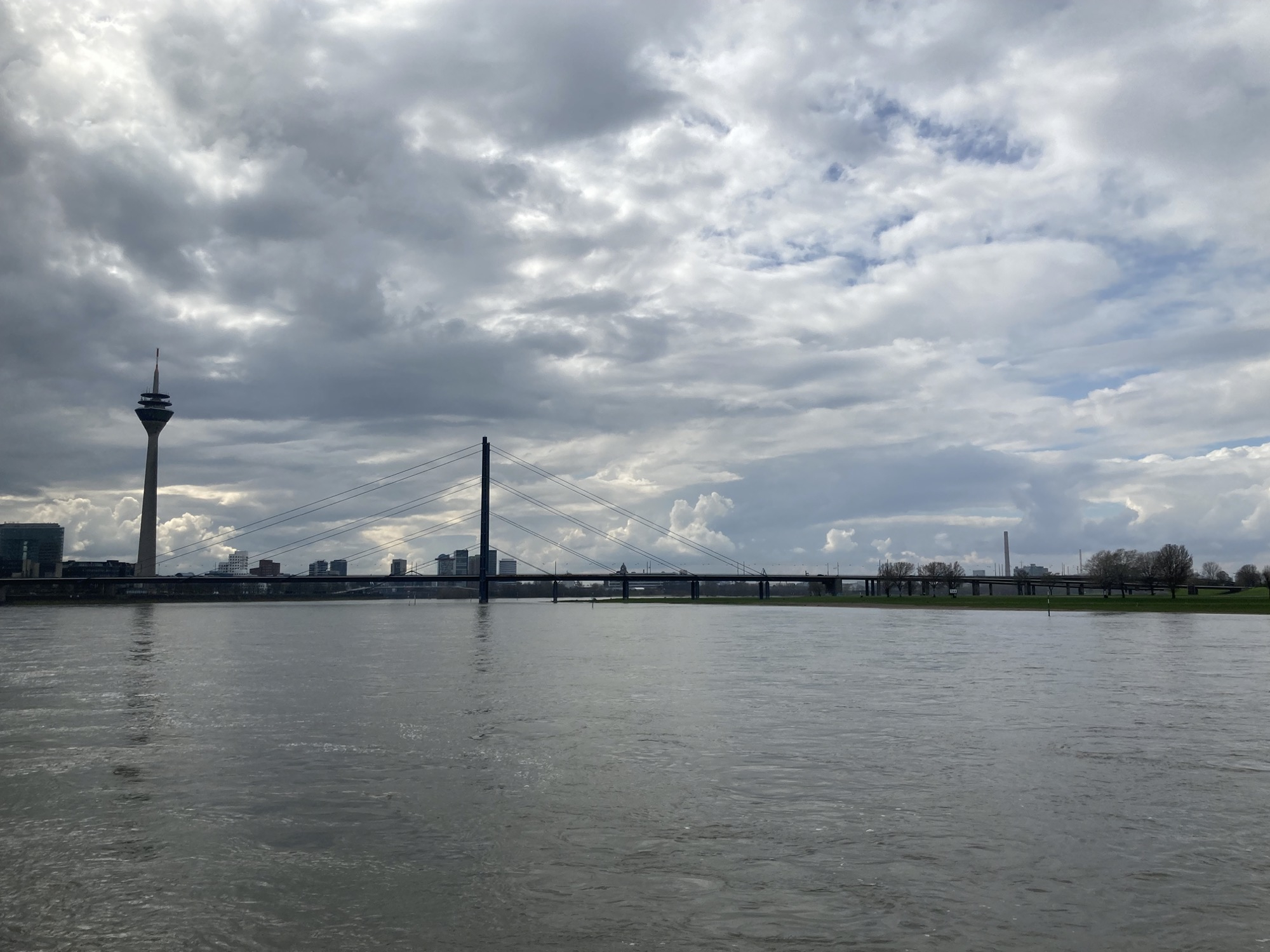 Blick von der Uferpromenade über den Rhein, im Wasser spiegeln sich ein Fernsehturm, eine Brücke sowie die aufreißenden Wolken am Himmel