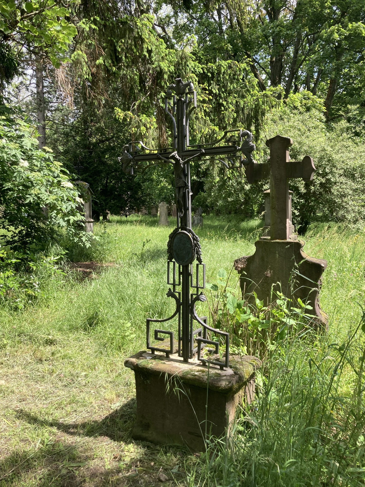 alte Grabsteine inmitten grüner Bäume, Sträucher und Gräser, der vordere Grabstein hat ein Metallkreuz, dahinter ragt ein Steinkreuz auf