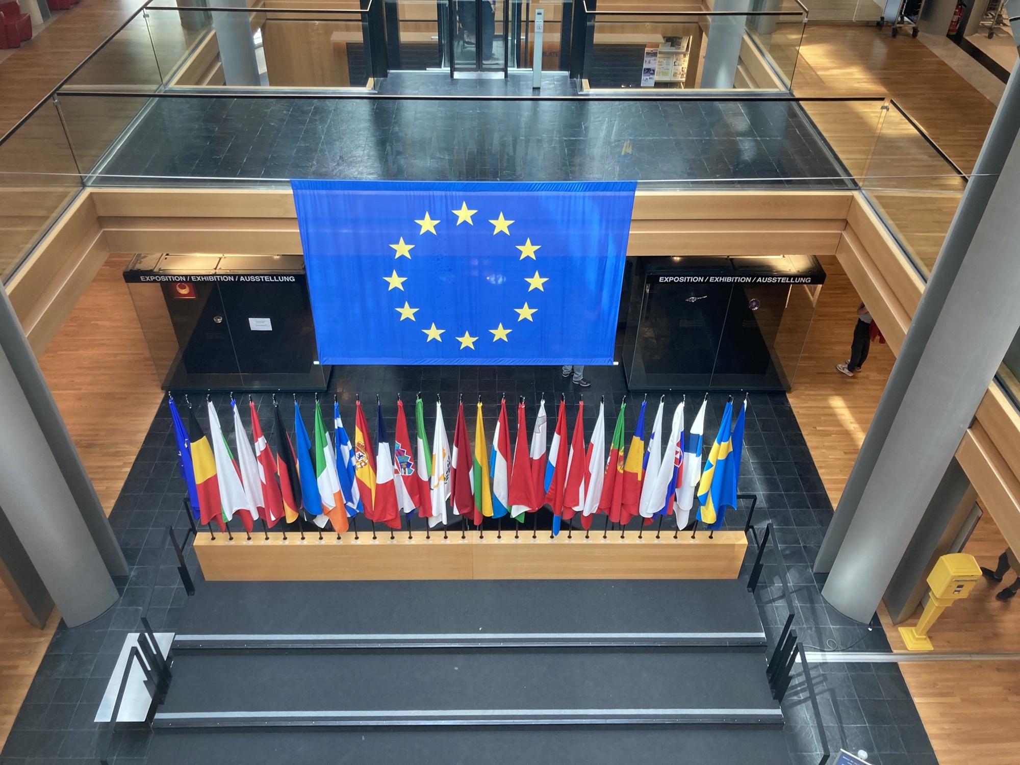 im europäischen Parlament, unter der großen EU-Flagge sind die Flaggen der Mitgliedsländer nebeneinander aufgereiht, rechts am Rand des Bildes ist ein gelber Postkasten zu sehen