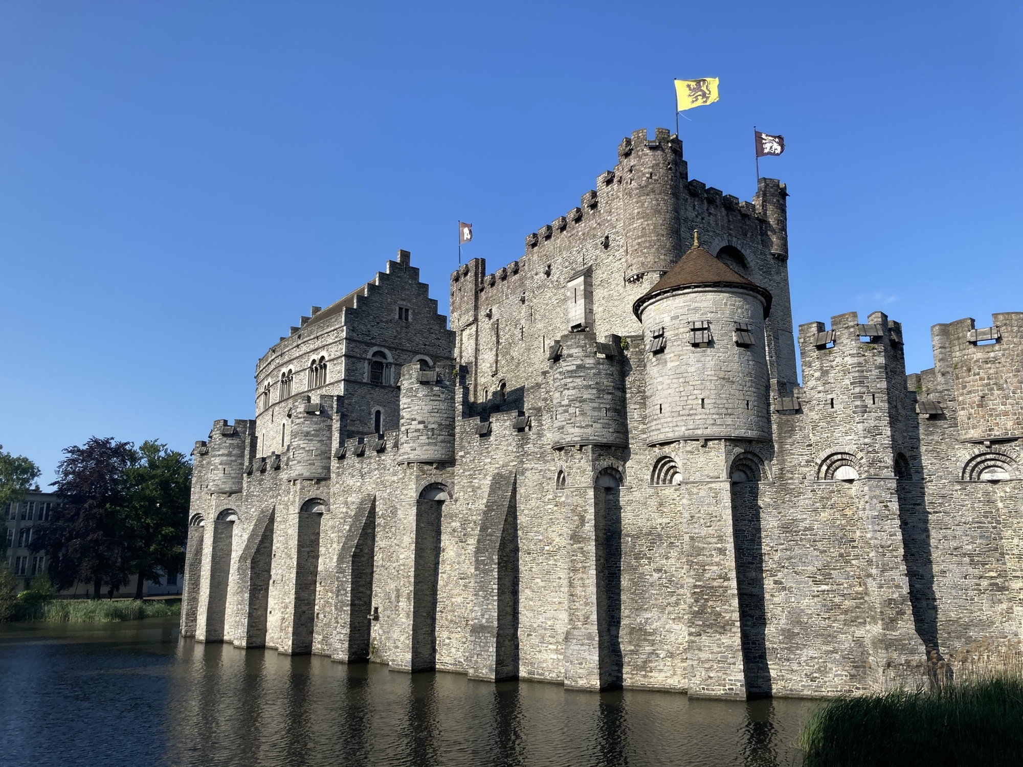 Burgmauern ragen aus dem Wasser empor, die Wände enden oben in Wachtürmen mit Schießscharten, auf dem Gebäude dahinter wehen gelbe und schwarze Flaggen, die das Stadtwappen zeigen