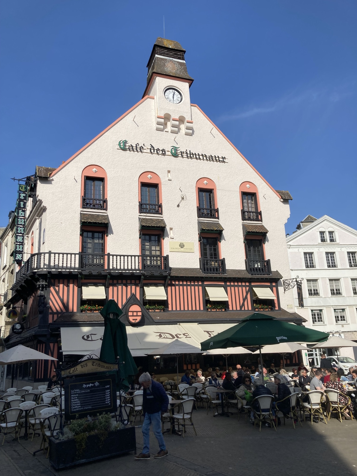 „Café de Tribunaux“, ein helles altmodisches Gebäude mit einem Turm mit Uhr oben drauf, der Gastgarten vor dem Haus liegt bereits zur Hälfte im Schatten