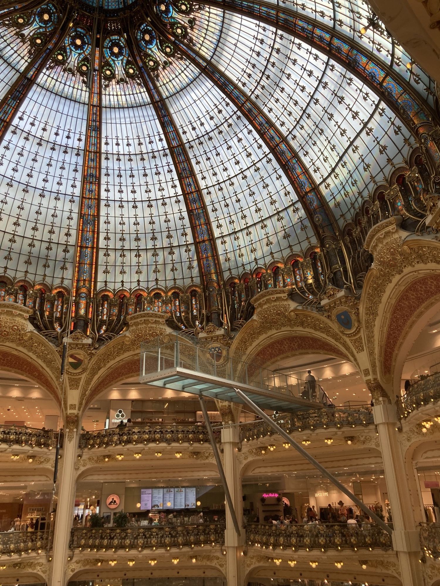 monumentales Glaskuppeldach im Inneren der Galeries Lafayette, zu sehen ist die opulente Glaskuppel und zwei Stockwerke darunter, vom obersten Stockwerk ragt ein Glasgang in den Kuppelbereich hinein