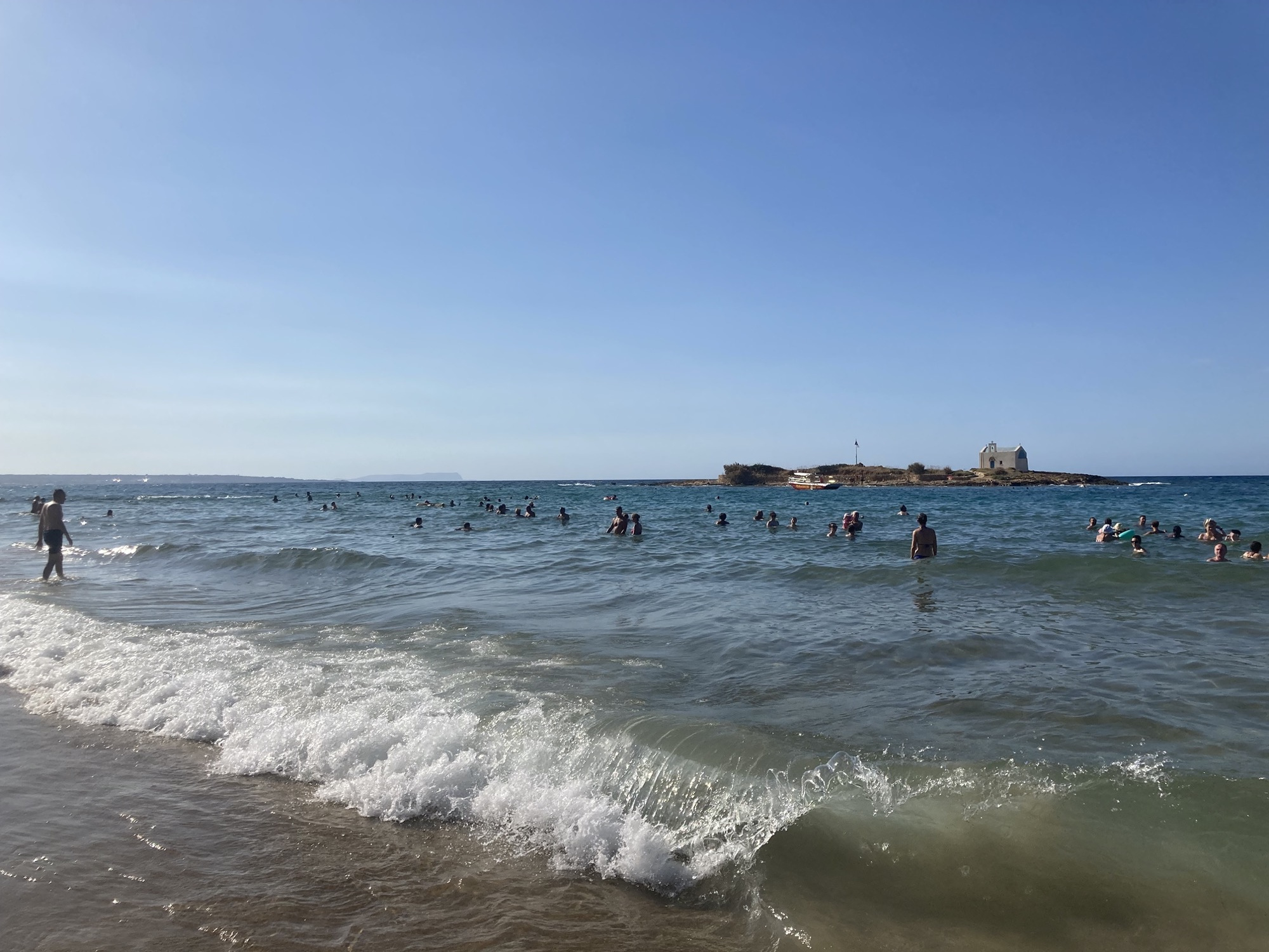 Strand von Malia, die Wellen brechen sich am Ufer, im seichten Wasser baden viele Menschen, im Hintergrund ist die Insel zu sehen, auf der sich eine kleine Kirche und ein Geocache befinden