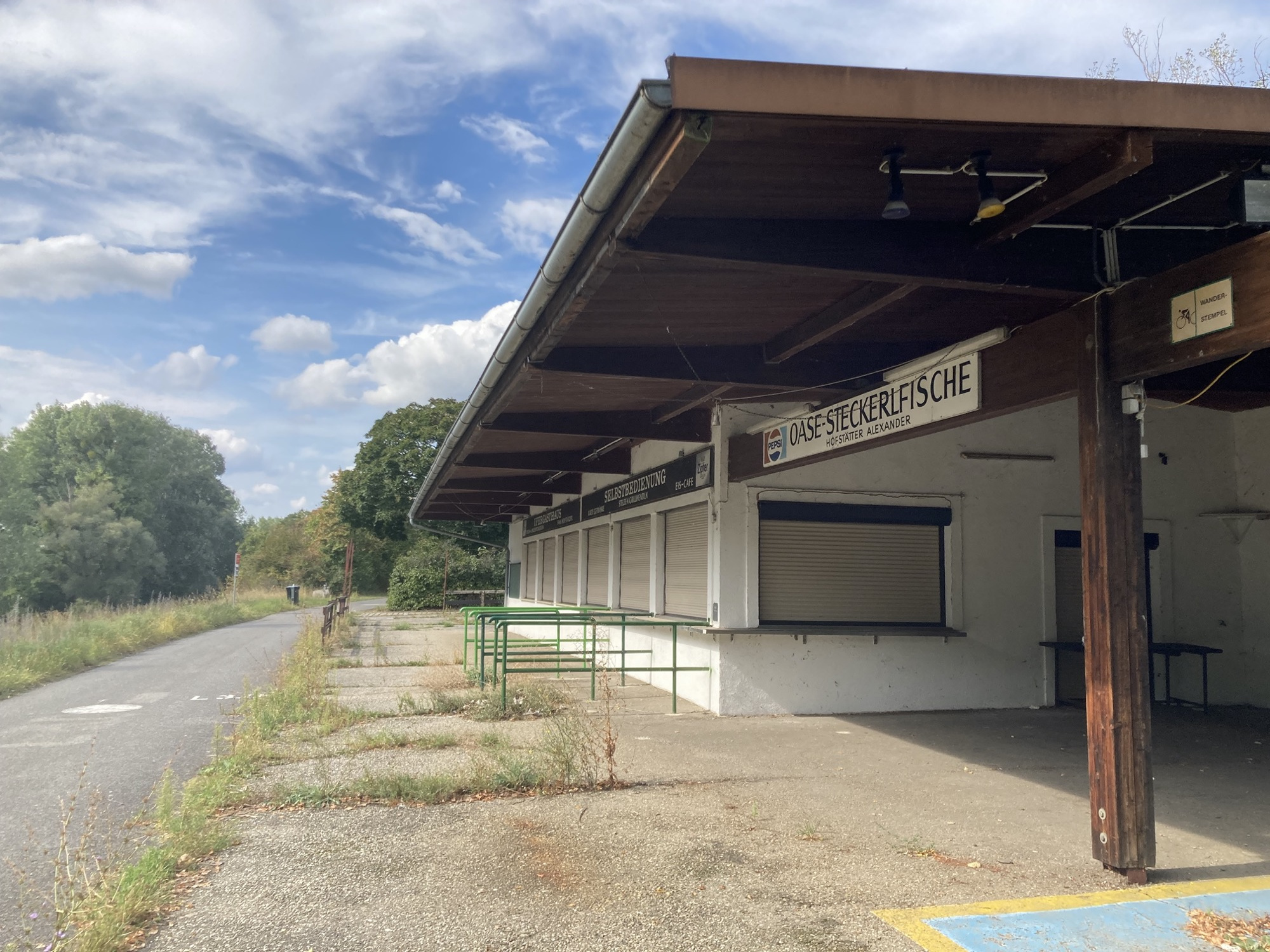 Ausflugslokal auf der Donauinsel, die Selbstbedienungsschalter sind mit Rolladen verschlossen, auf einem Schild steht Oase-Steckerlfische, Hofstätter Alexander“, der Asphalt vor dem Lokal ist von Pflanzen durchbrochen