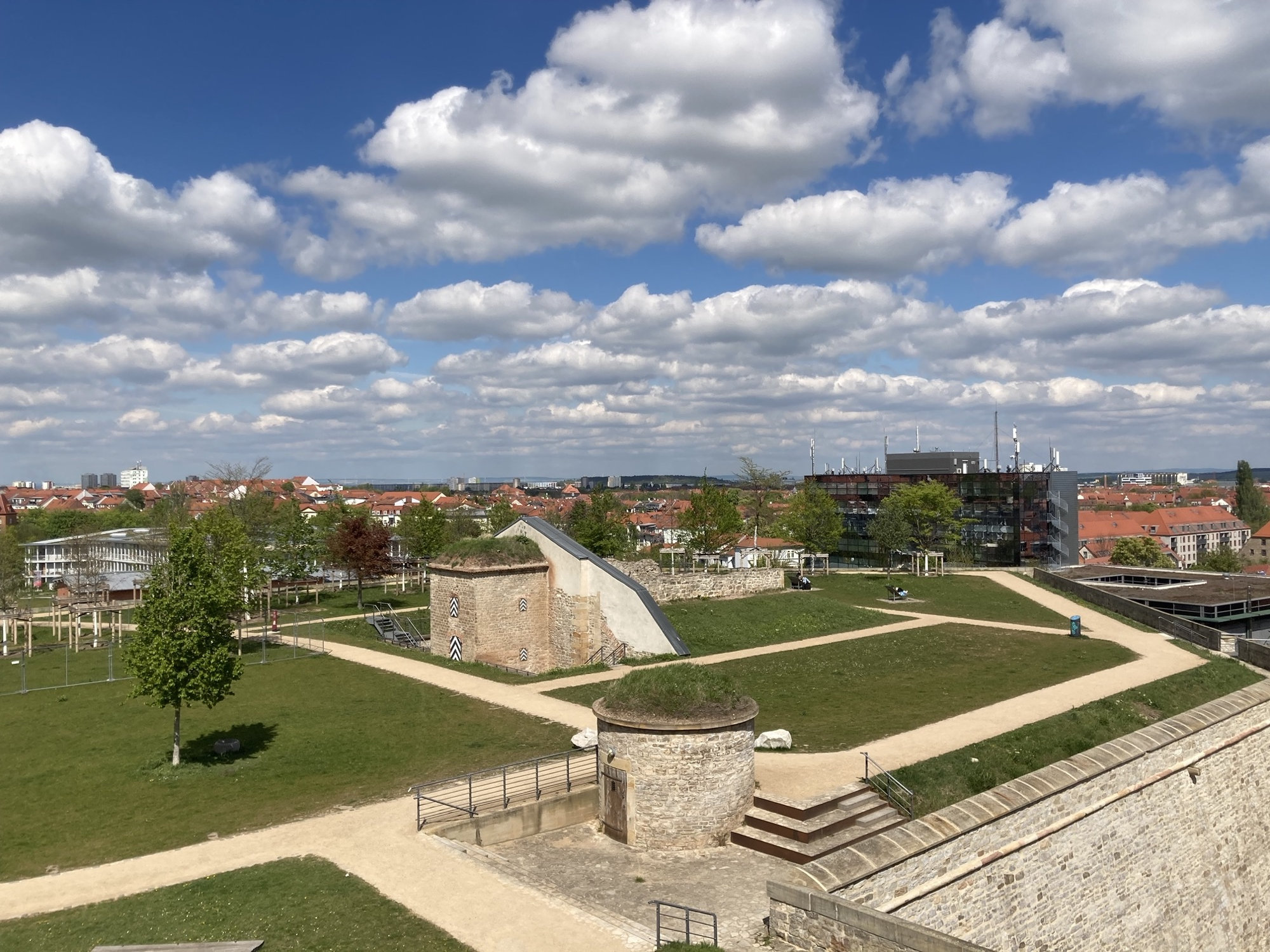Überblick über die Citadelle Petersberg, Grünflächen kombiniert mit alten Festungsanlagen, in der Mitte ein ehemaliges Munitionslager, am blauen Himmel viele weiße Wölkchen
