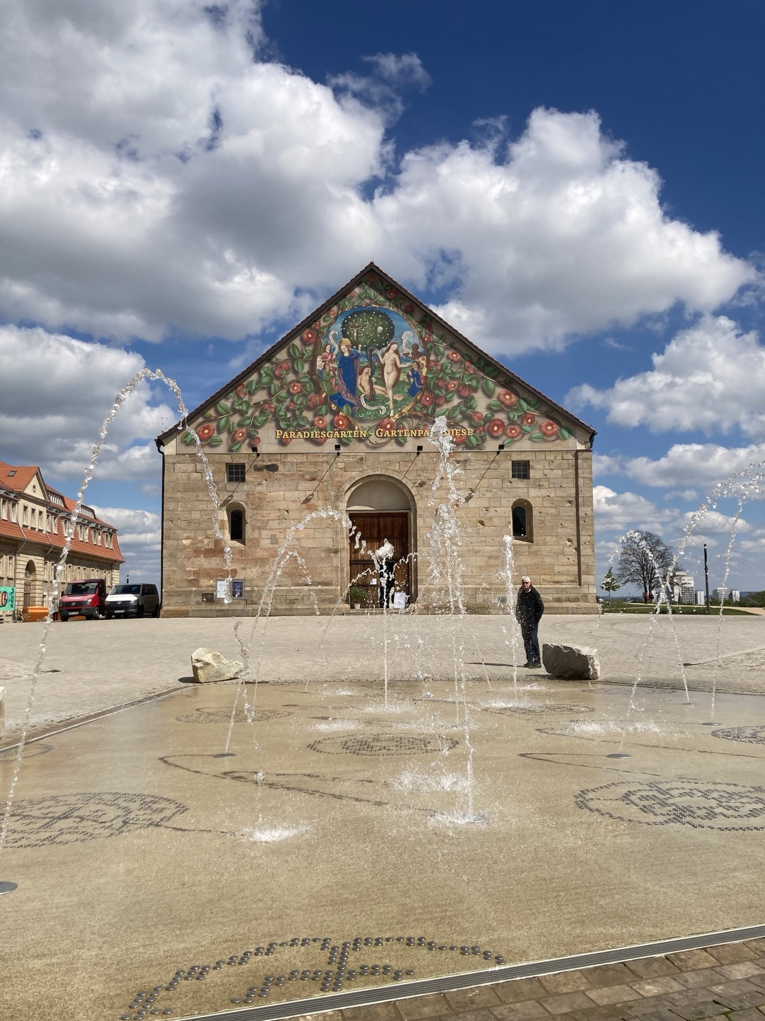 Schmalseite der Peterskirche auf dem Petersberg, im dreieckigen Dachbereich wird eine Gartenausstellung mit einem Paradiesbild beworben, davor ein ebenerdiger Springbrunnen mit Wasserfontainen