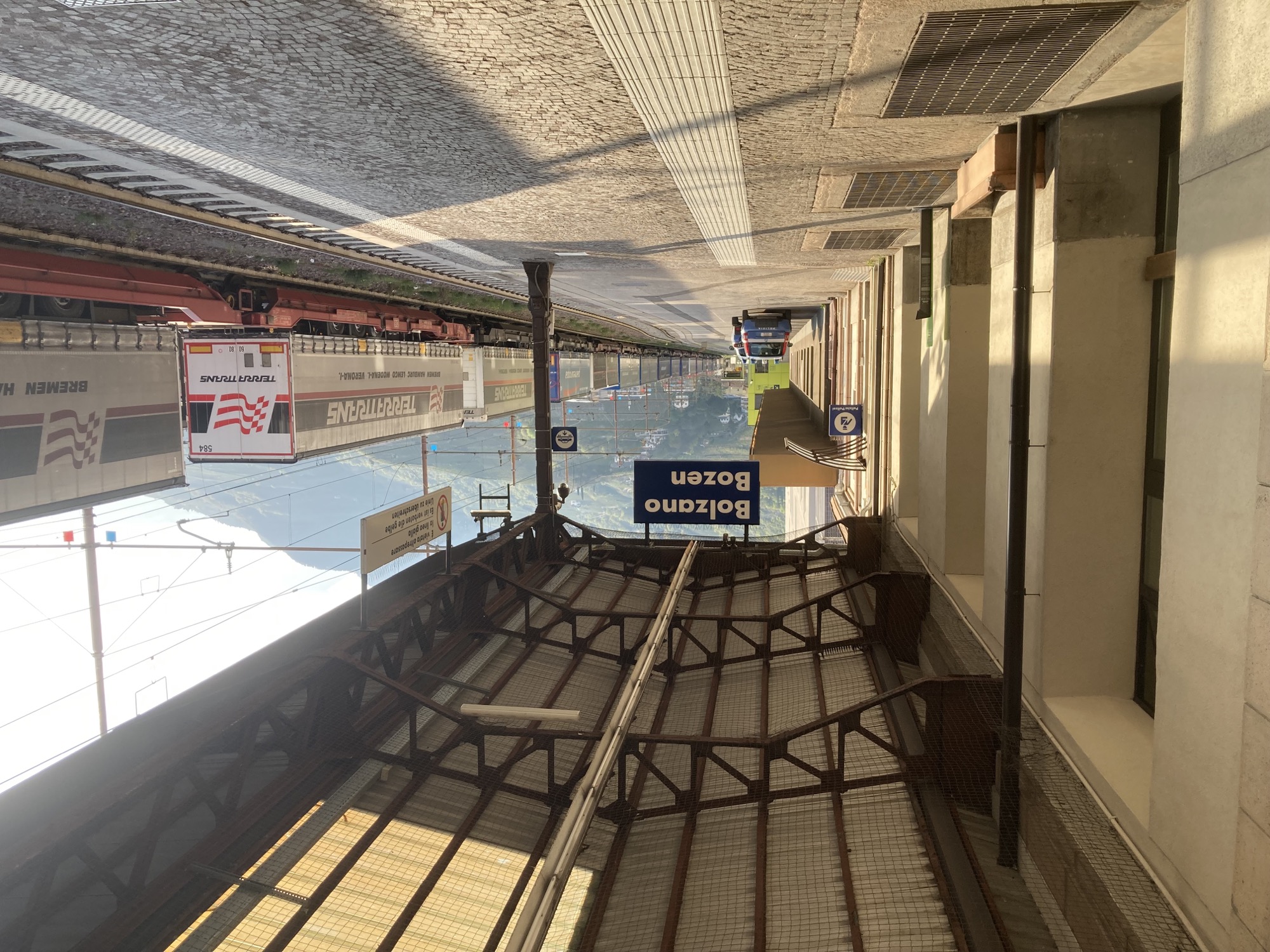 Bahnsteig 1 am Bahnhof Bolzano/Bozen, von rechts strahlt Morgenlicht ins Bild, die Frachtwaggons auf Gleis 2 werfen lange Schatten