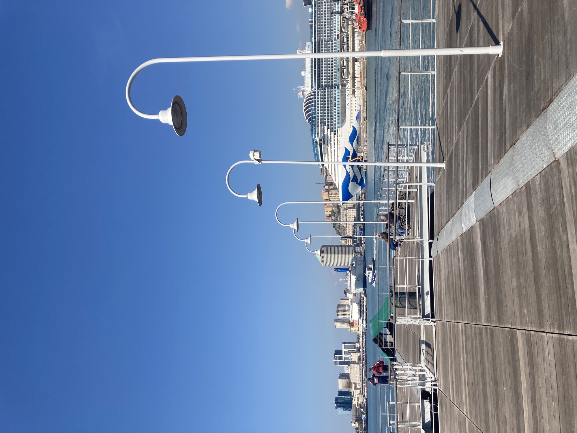 auf dem Meer schwimmende Holzplattform mit weißem Geländer, Sitzbänken und weißen Lampen, im Hintergrund ein großes Kreuzfahrtschiff