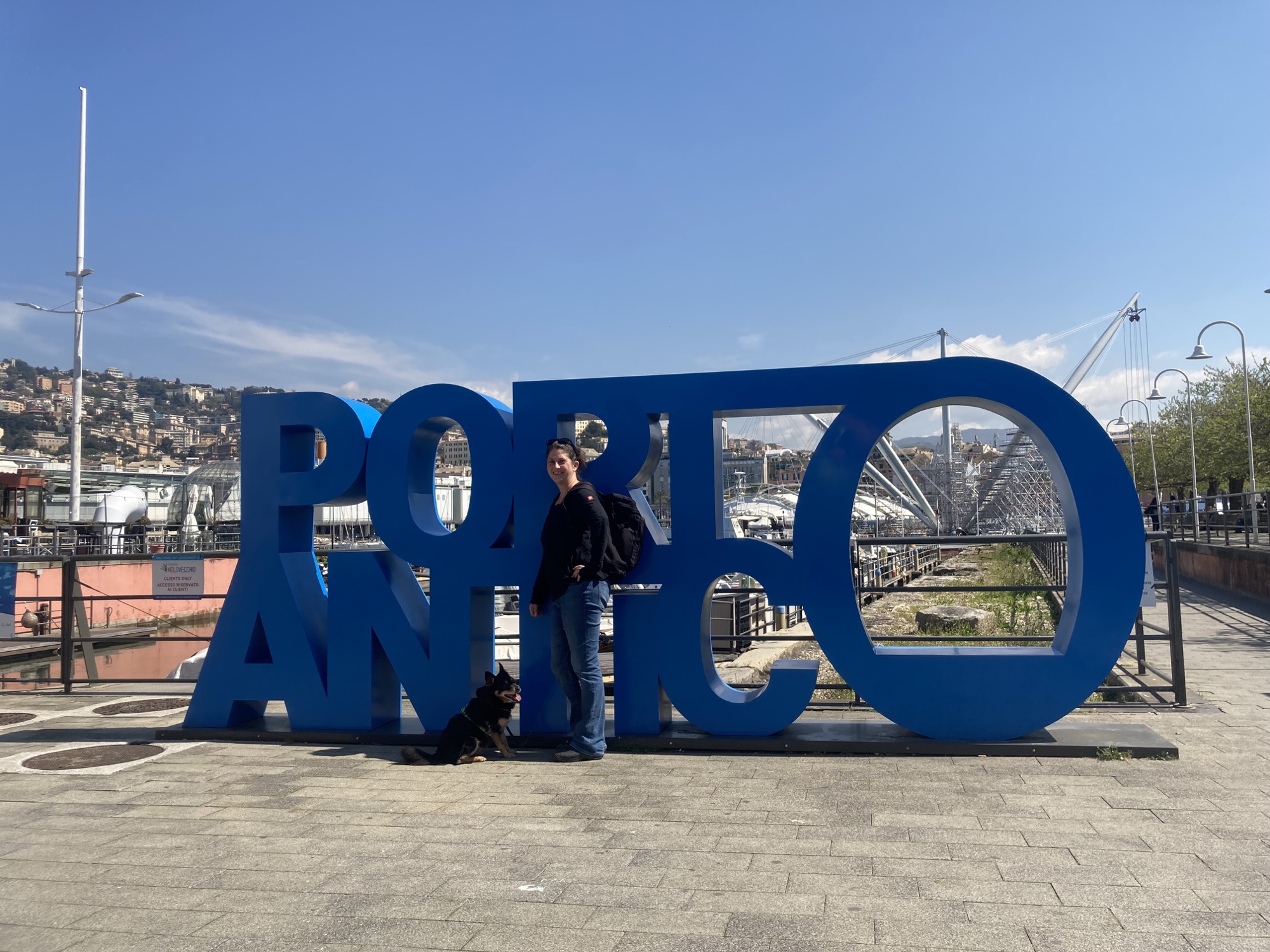 menschengroßer blauer Schriftzug „Porto Antico“, mittig davor steht eine Person in Jeans, schwarzem Shirt und mit Rucksack, daneben ein kleiner schwarzbrauner Hund