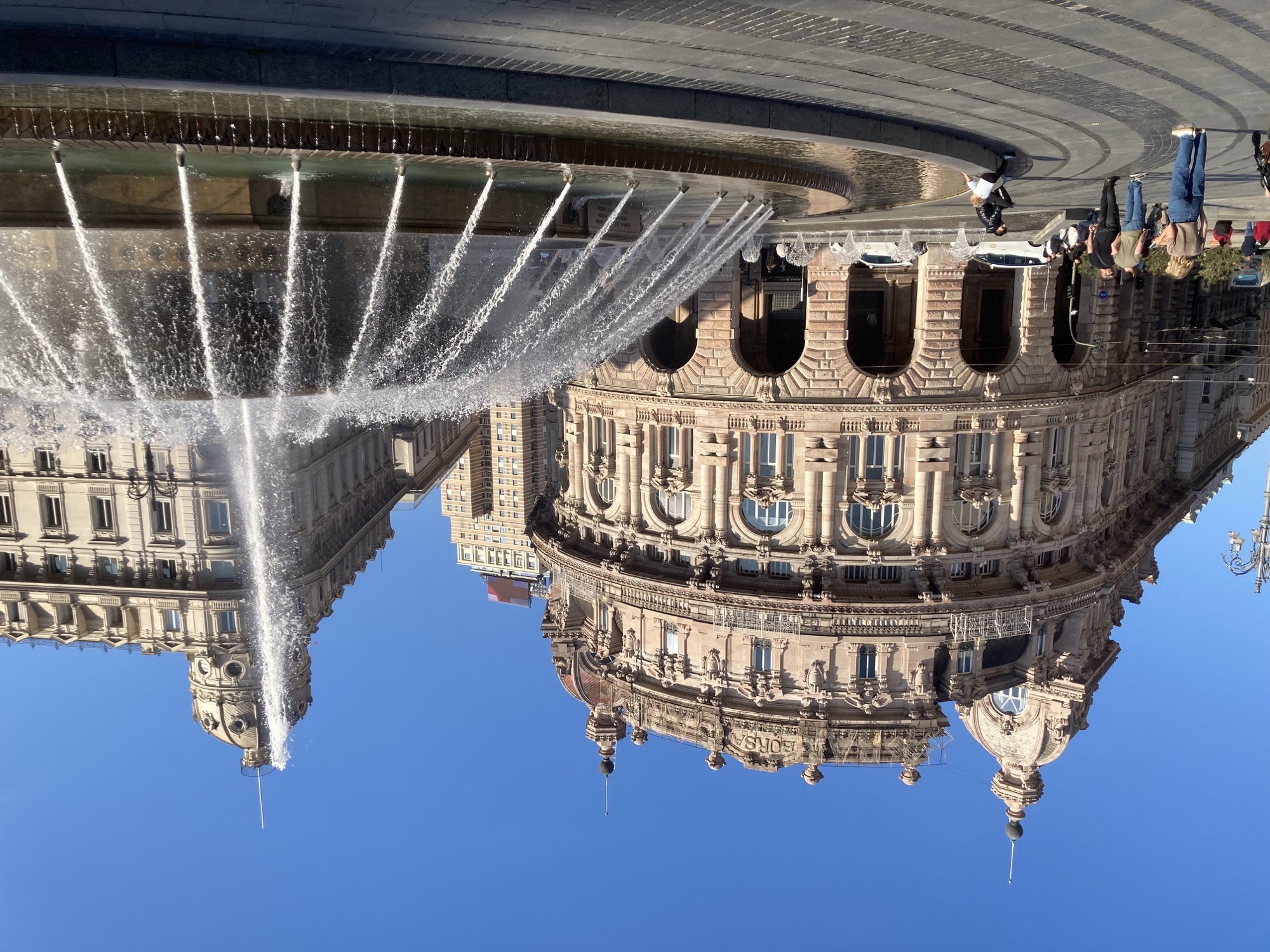 Springbrunnens auf der Piazza De Ferrari, rechts sprühen Fontänen auf den runden Mittelteil des Brunnes, im Hintergrund ein dekoratives Gebäude mit vielen Säulen und Verzierungen, ganz oben ist der Schriftzug „Borsa“ zu lesen