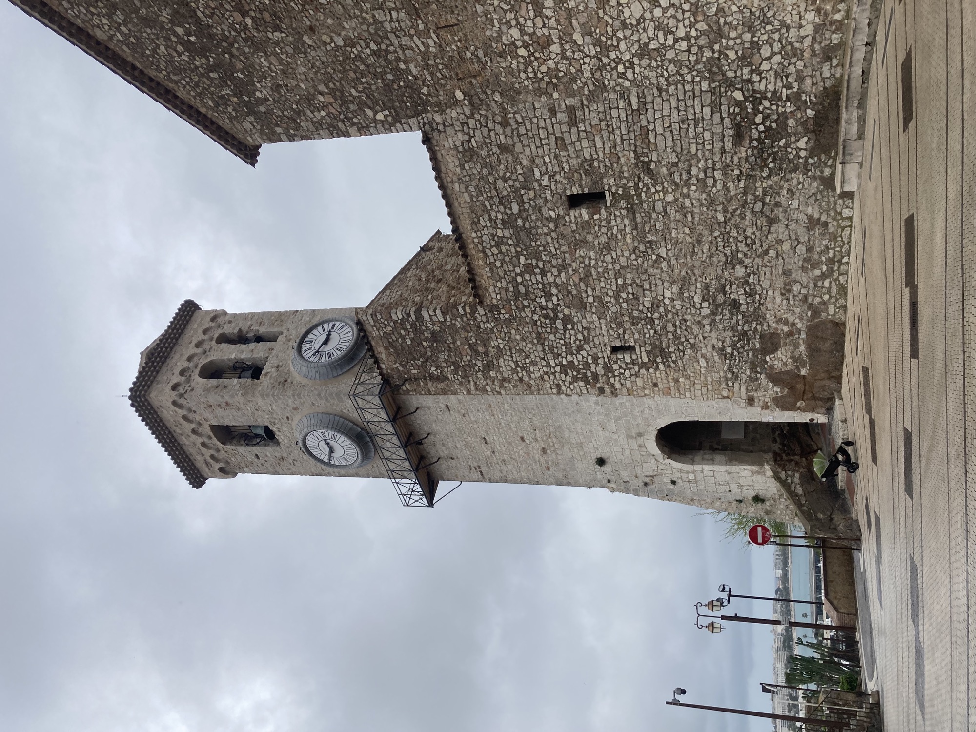 Kirchturm der romanischen Kirche Notre-Dame-d'Espérance de Cannes, grauer bewölkter Himmel, der Boden ist leicht feucht, den Turm ziert eine Turmuhr mit römischen Ziffern, sie zeigt etwa 10 Minuten vor 3 an