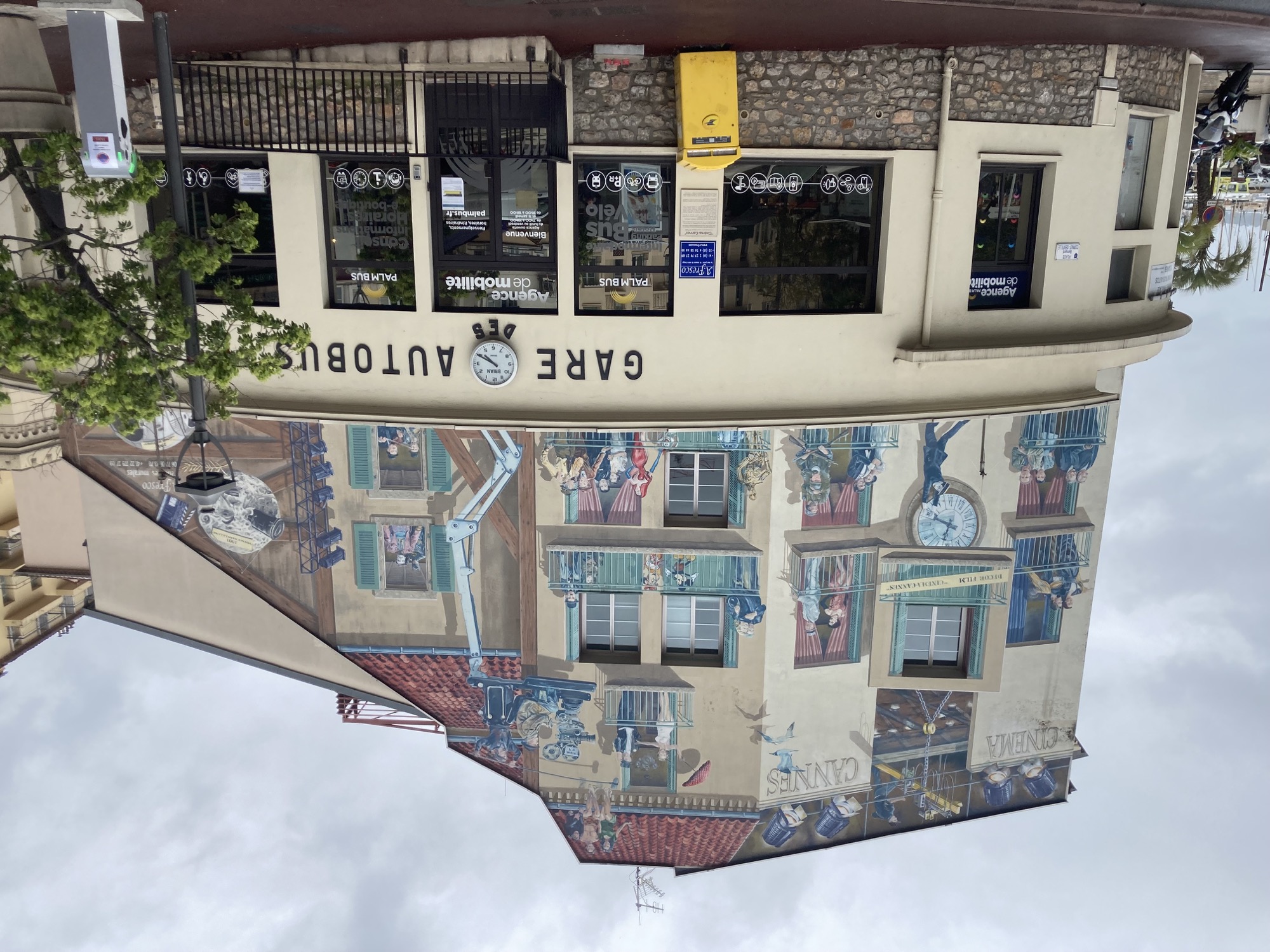 Gare Autobus in Cannes, ein Gebäude mit großen Fenstern im Erdgeschoss, darüber ist die Wand mit einem detailreichen Graffiti bemalt, zwischen den realen Fenstern sind Fenster und Balkone mit bekannten Filmfiguren aufgemalt zB Jack und Rose aus Titanic und Batman und den Joker