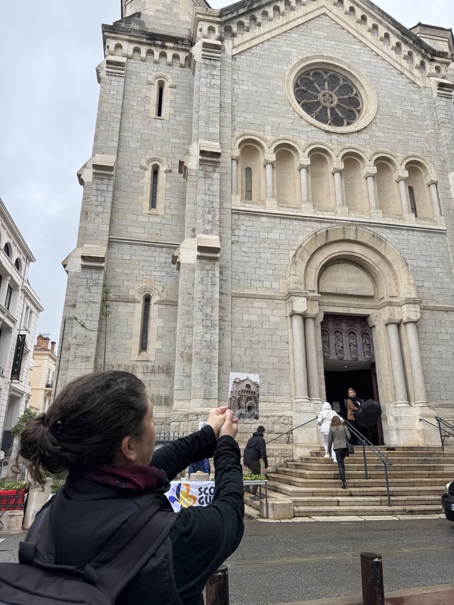 Front der neoromanischen Kirche Notre-Dame-de-Bon-Voyage, links unten/vorne im Bild steht eine Person und hält in beiden Händen ein Foto, das genau dieselbe Kirchenfront zeigt