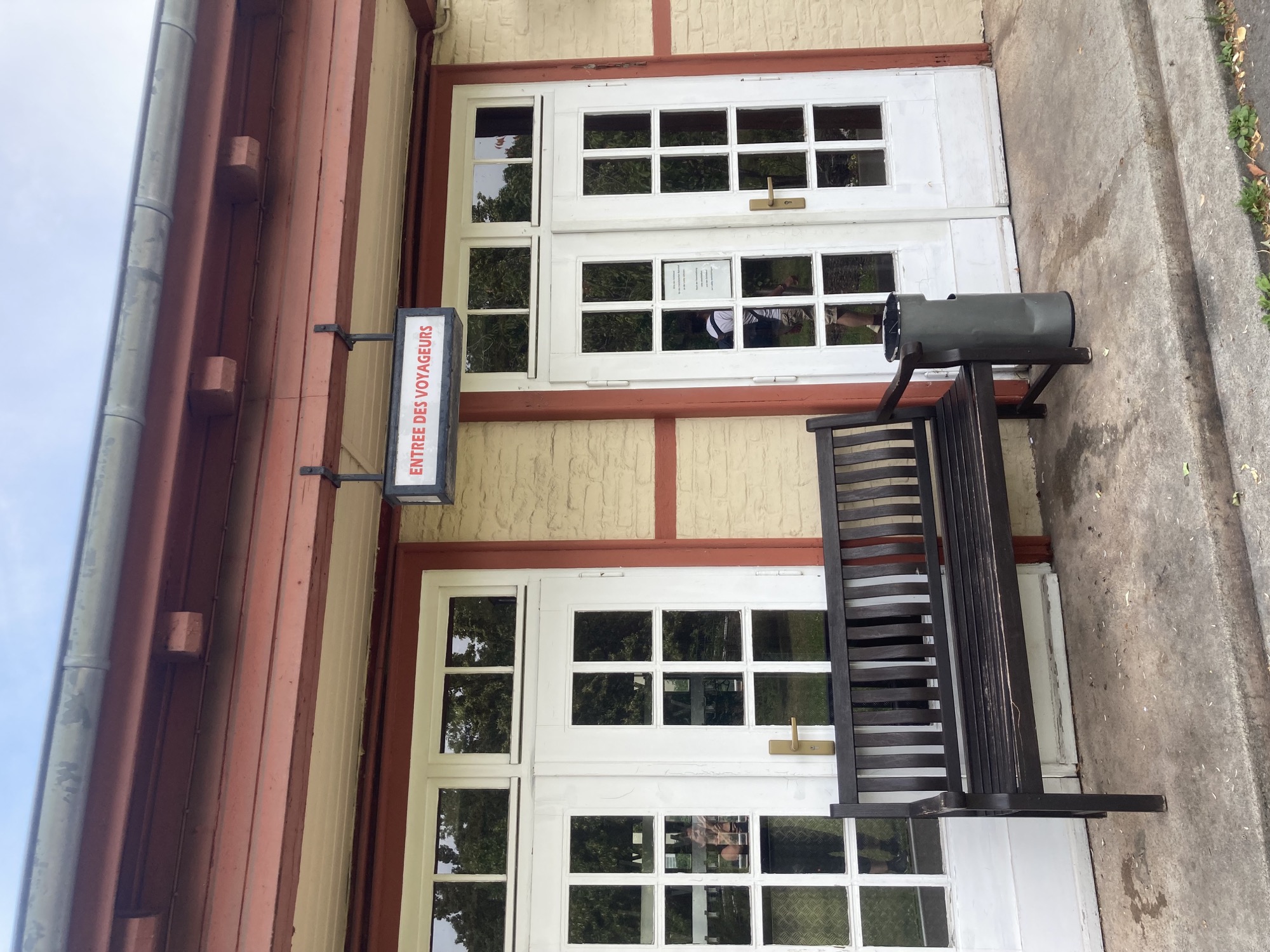 einstöckiges Gebäude mit zwei weißen Doppelflügeleingangstüren, vor einer Tür steht eine Holzbank und daneben ein Mistkübel, darüber hängt ein weißes Schild mit roter Schrift „Entree des Voyageurs“