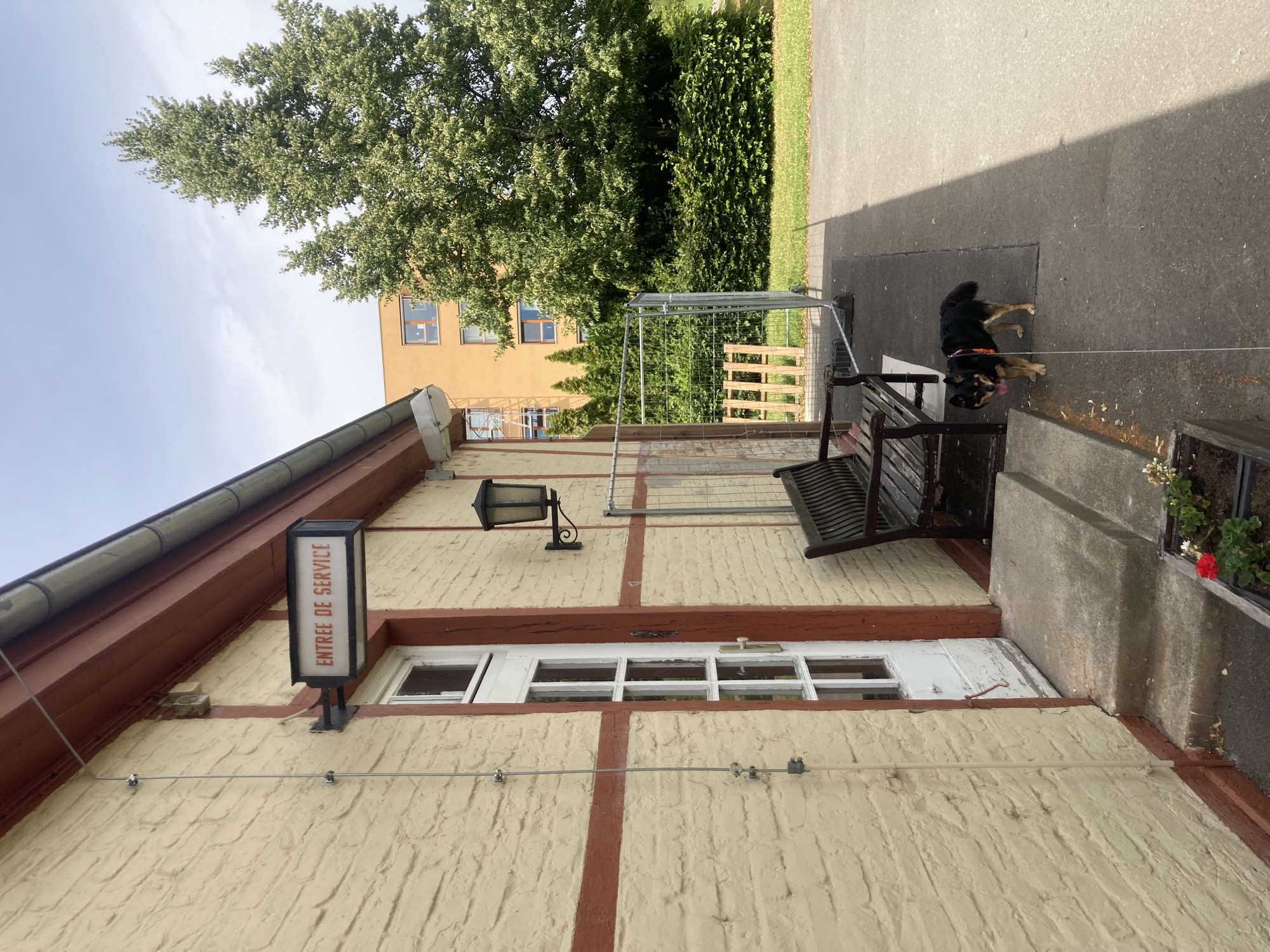 Hochformat desselben Gebäudes, hier mit einer normalen weißen Tür, darüber ein Schild „Entree de Service“, vor der Tür im unteren Bereich des Bilds ein kleiner schwarzbrauner Hund