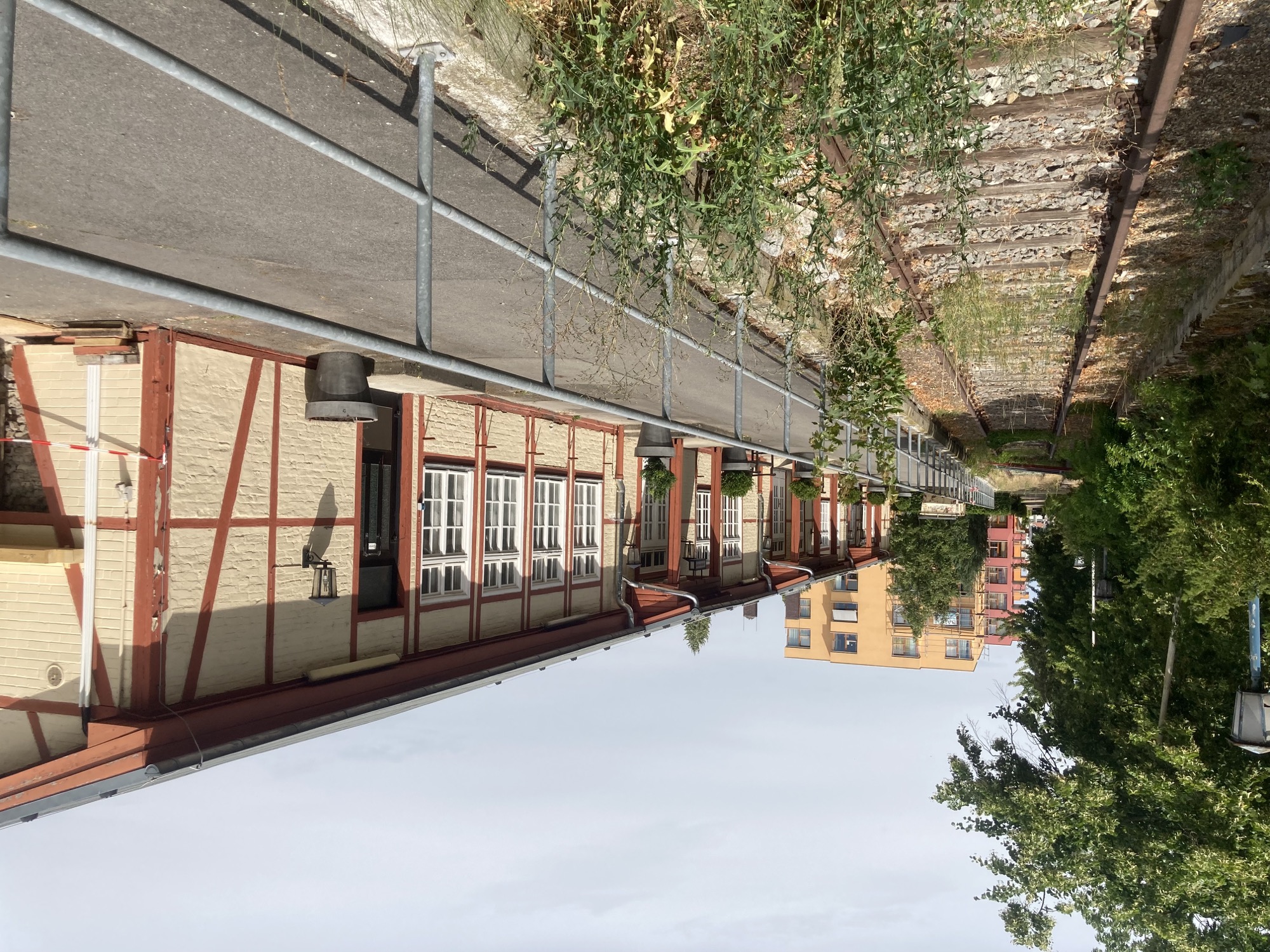 einstöckiges, langgestrecktes Gebäude, aus Ziegeln erbaut, die Mauern sind weiß gestrichen und mit braunen Balkenzeichnungen verziert, links im Bild ein nicht mehr genutztes Bahngleis (viel Unkraut wächst zwischen den Schienen)