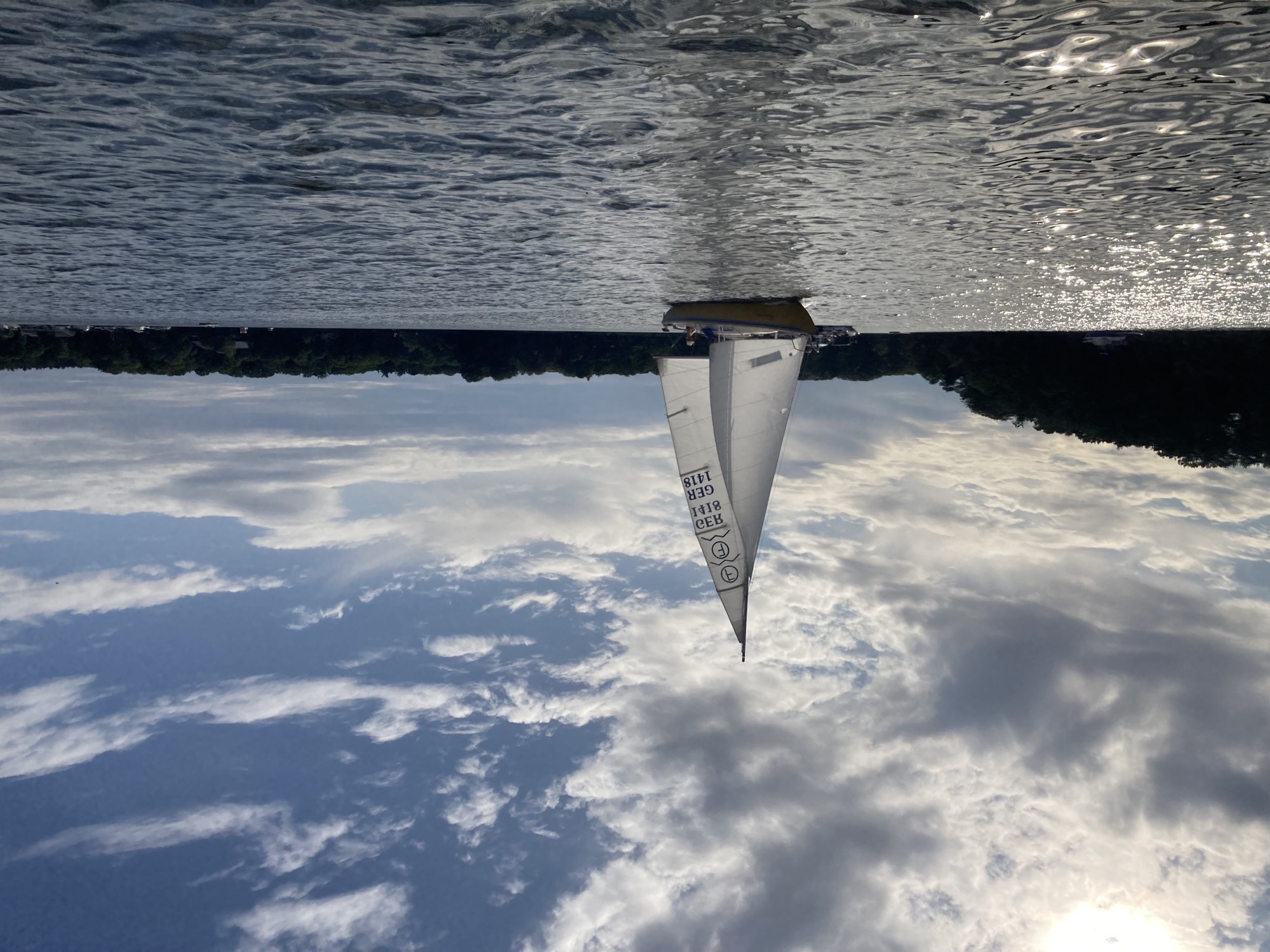 Foto eines Segelboots mit zwei aufgespannten weißen Segeln auf einem glitzernden See mit leicht bewölktem, blauem Himmel im Hintergrund