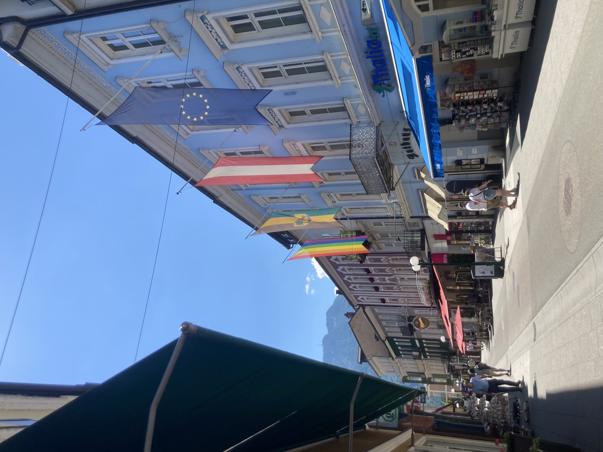 Fußgängerzone in strahlendem Sonnenschein, vor dem blauen Haus rechts im Bild hängen Flaggen (EU, Österreich, Bad Ischl und Regenbogen), alle Geschäfte sind von Markisen geschattet