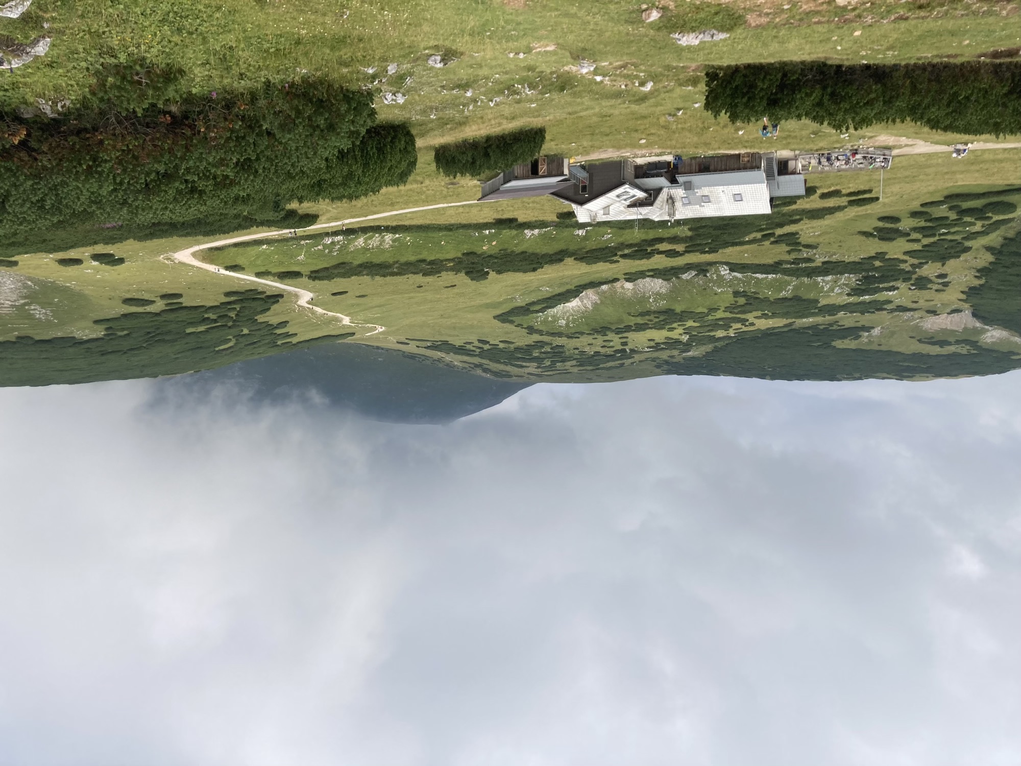 Berghütte Damböckhaus inmitten grüner, teils felsiger Hügel, rechts führt ein Wanderweg in Richtung des wolkenverhangenen Gipfels