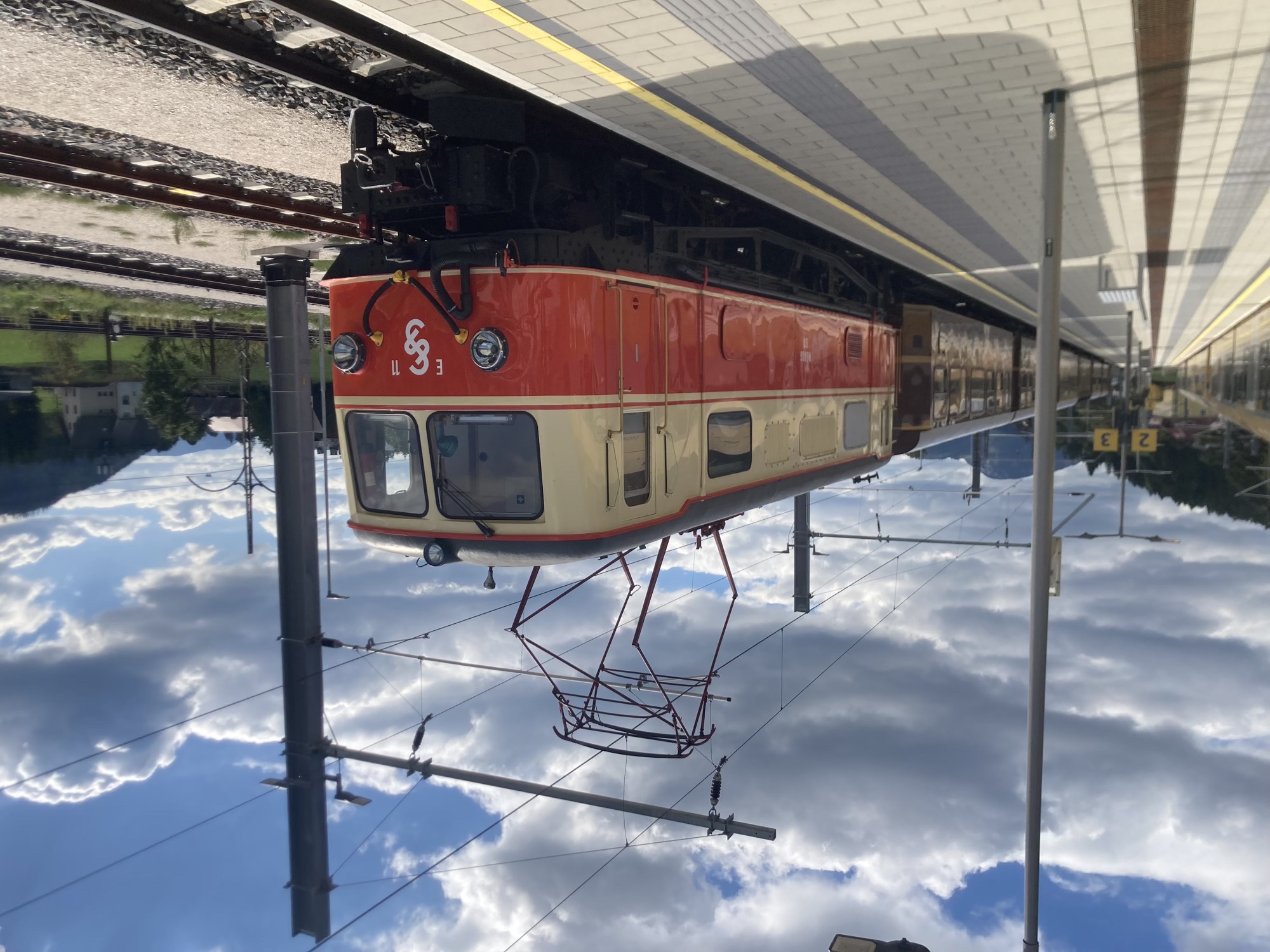 E-Lokomotive am Bahnsteig, vorne neben dein Scheinwerfern ein geschwungenes Zeichen und die Aufschrift E11, der untere Teil der Lokomotive ist rot gestrichen, der obere Teil beige